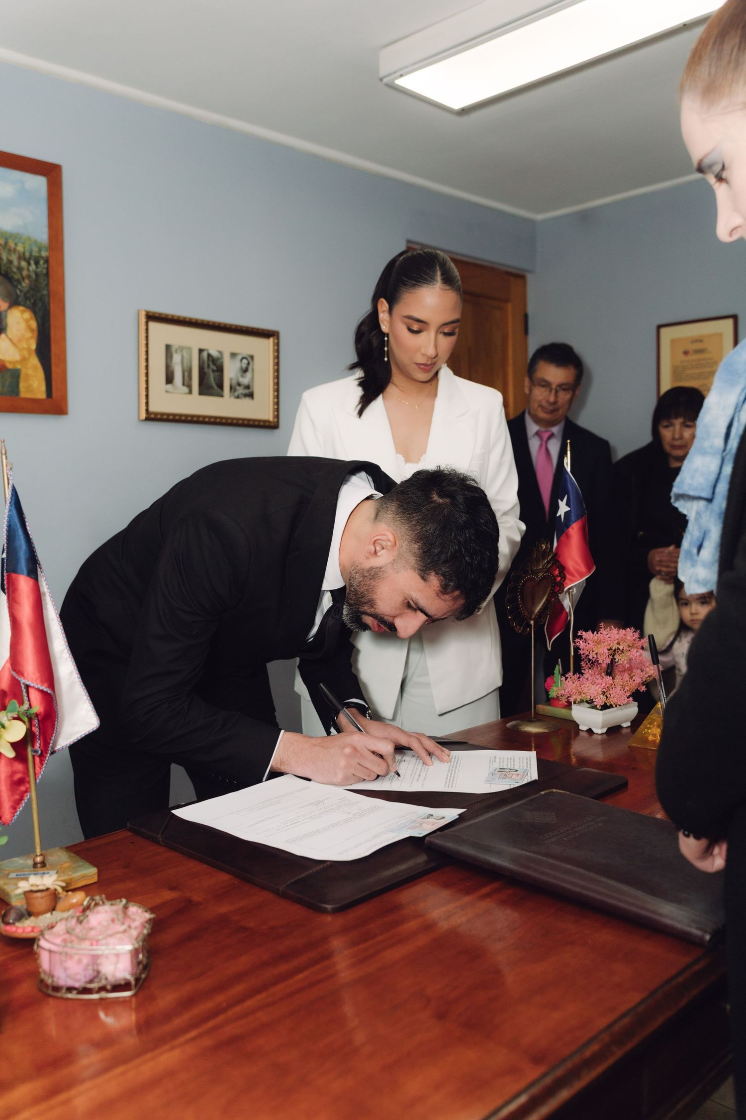 Groom in black suit signing wedding documents at a wooden desk during a civil ceremony.