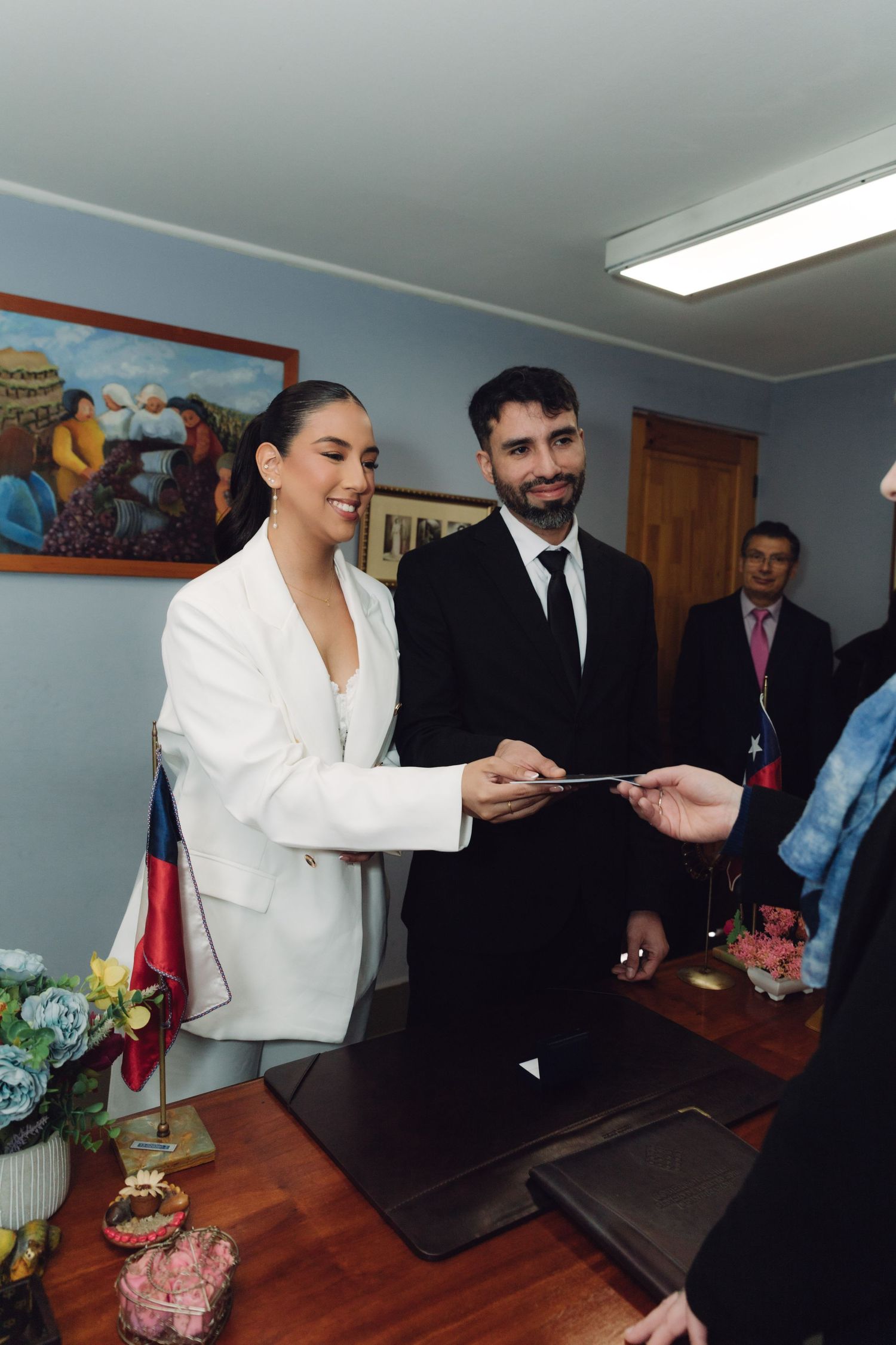 Smiling bride in white suit and groom in black suit receiving wedding documents.