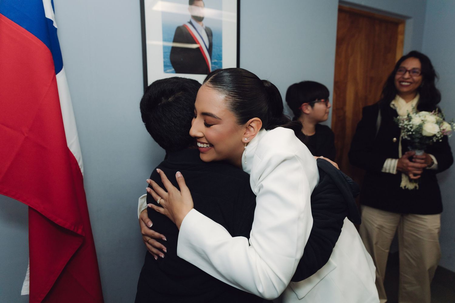 Smiling woman in white blazer hugging a guest near Chilean flag at an official civil ceremony.