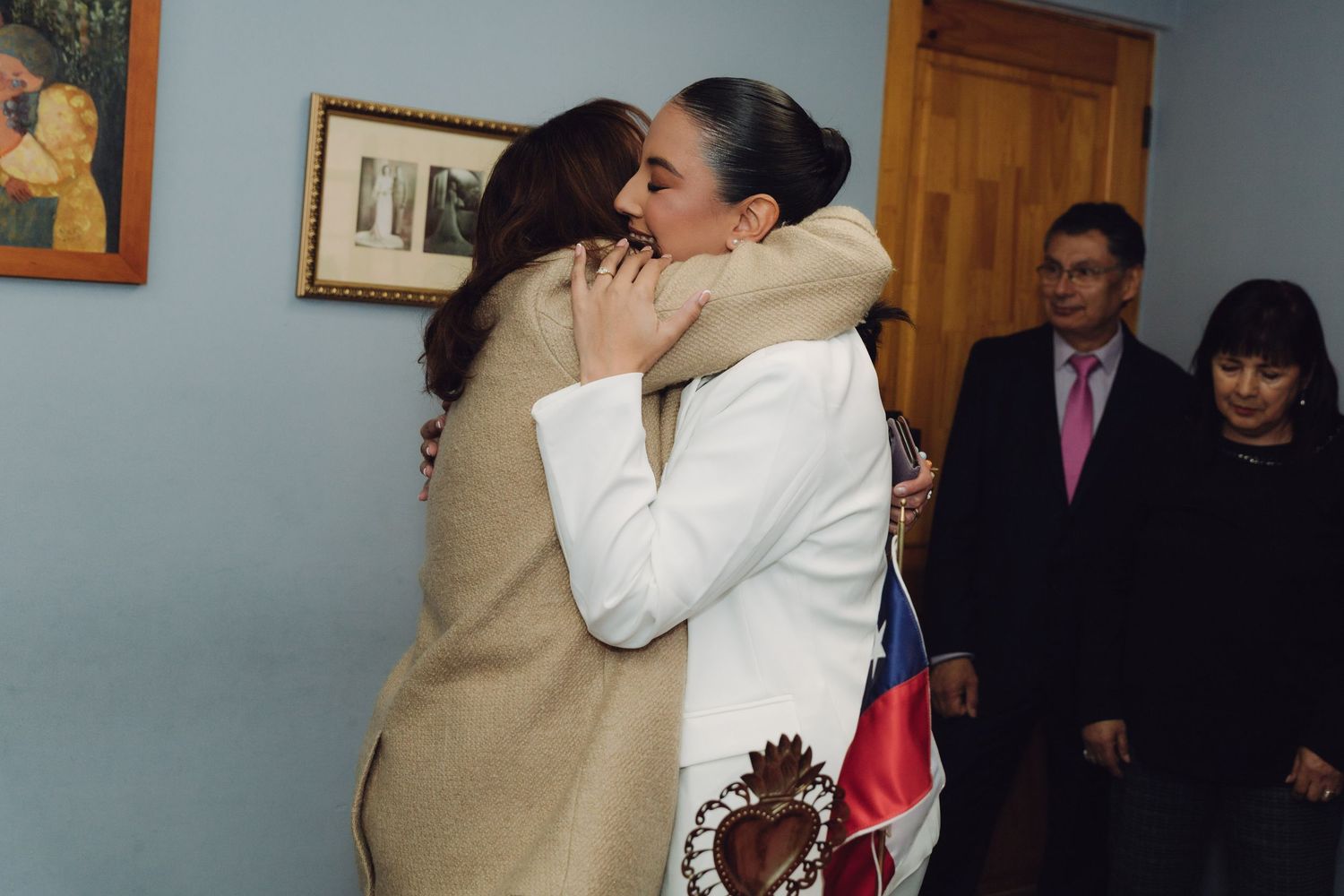 Two women sharing an emotional embrace at a Chilean civil ceremony while suited guests watch nearby.