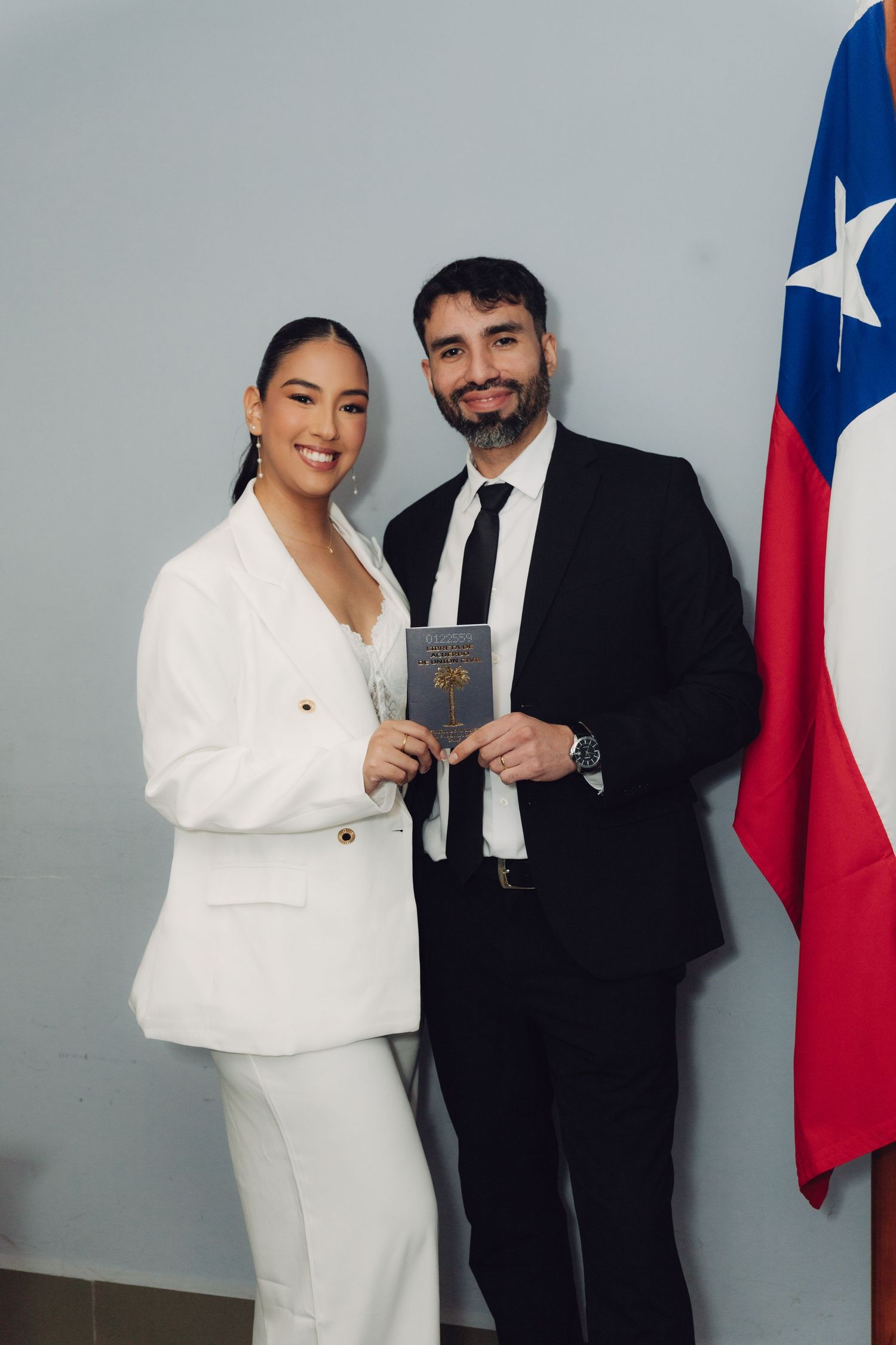 Couple in formal attire posing with official document beside Chilean flag at civil registry.