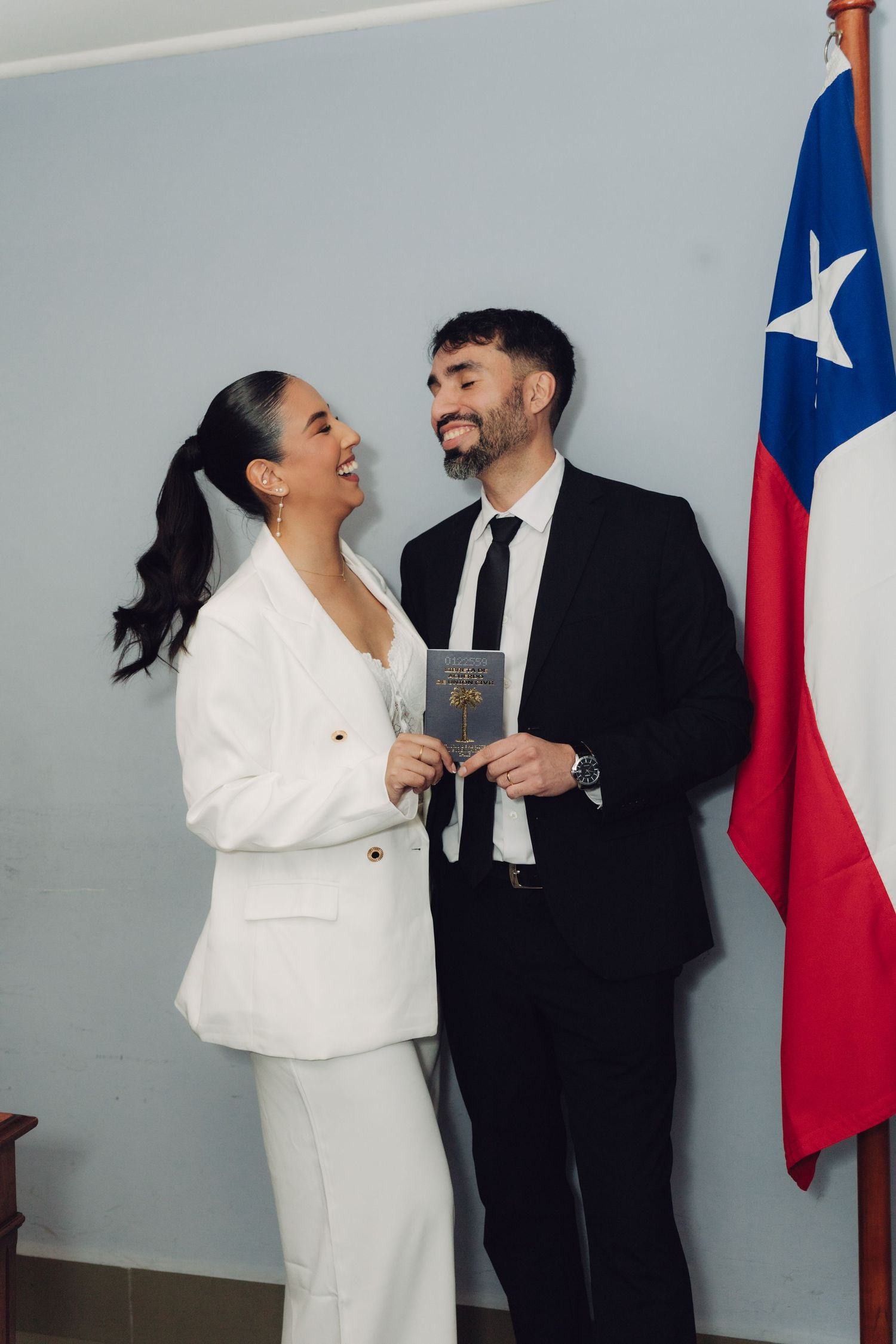 Happy couple in formal wear laughing together holding official document next to Chilean flag.