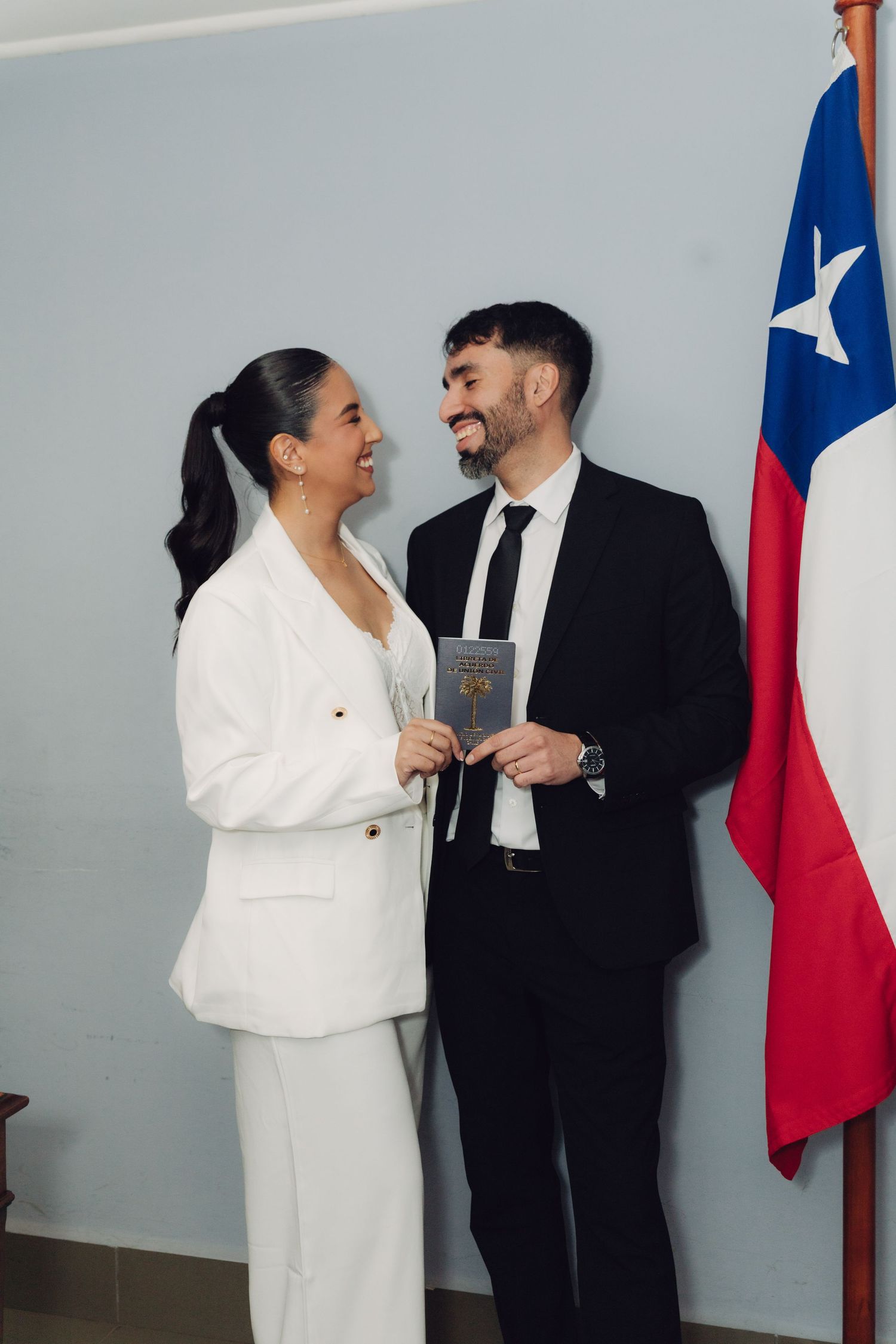 Newlywed couple smiling at each other holding marriage certificate beside Chilean national flag.