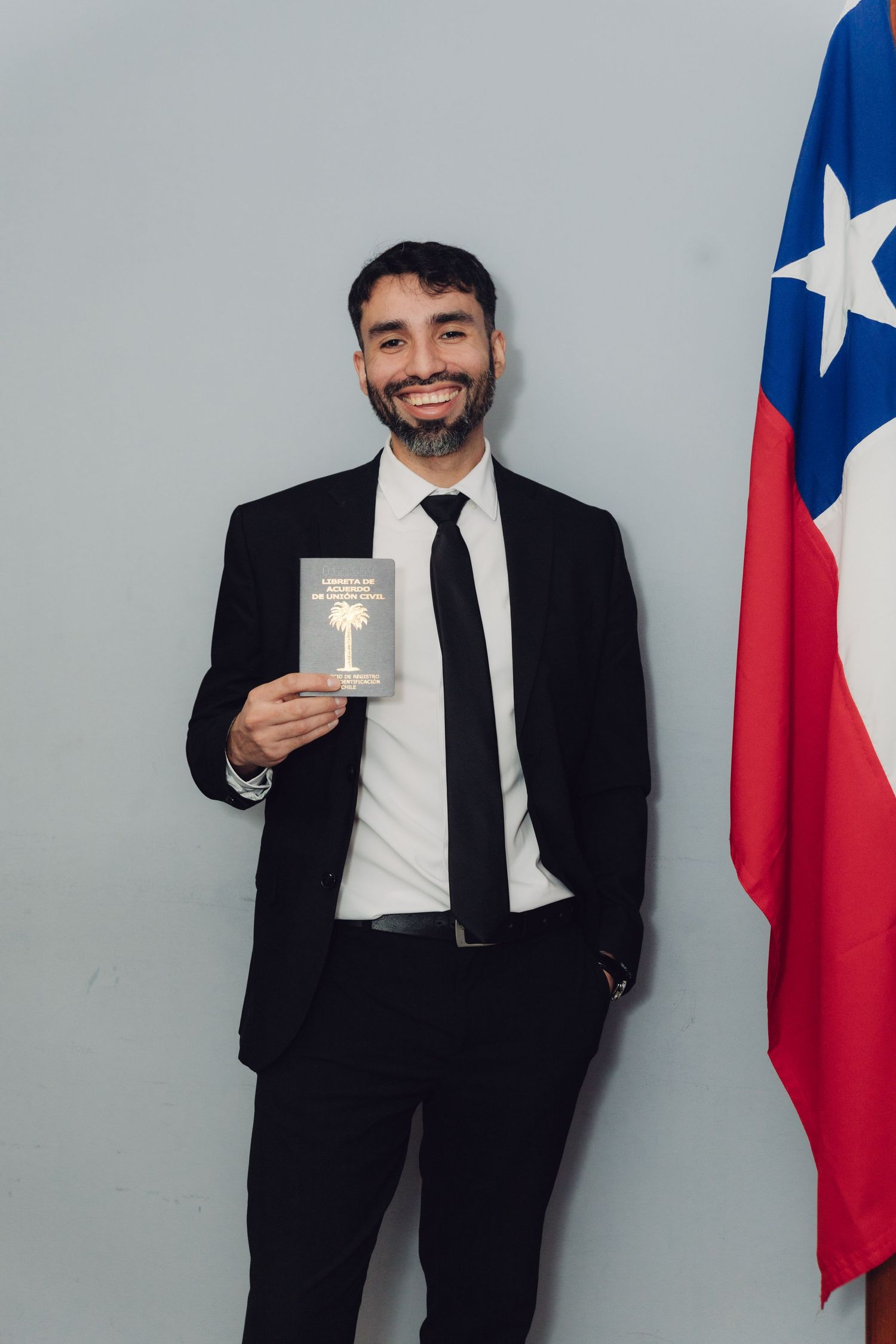 Smiling bearded man in black suit proudly displaying official Chilean document beside national flag.