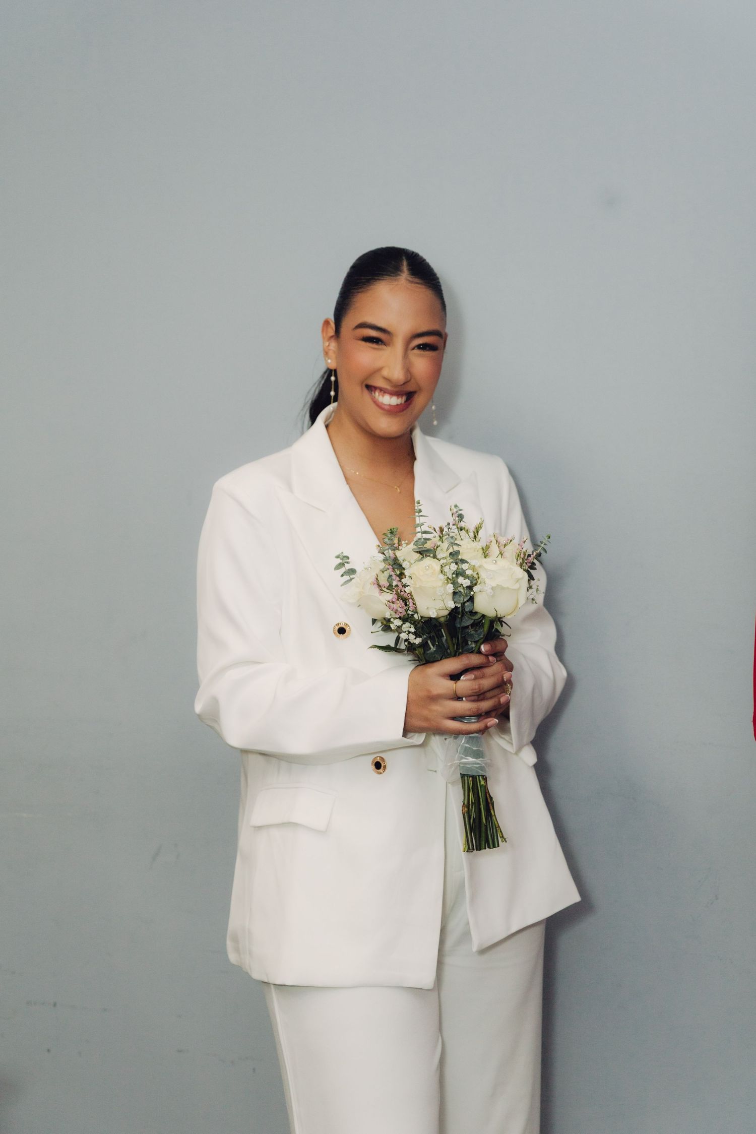 Smiling bride in white blazer suit holding bouquet of white roses against gray background.