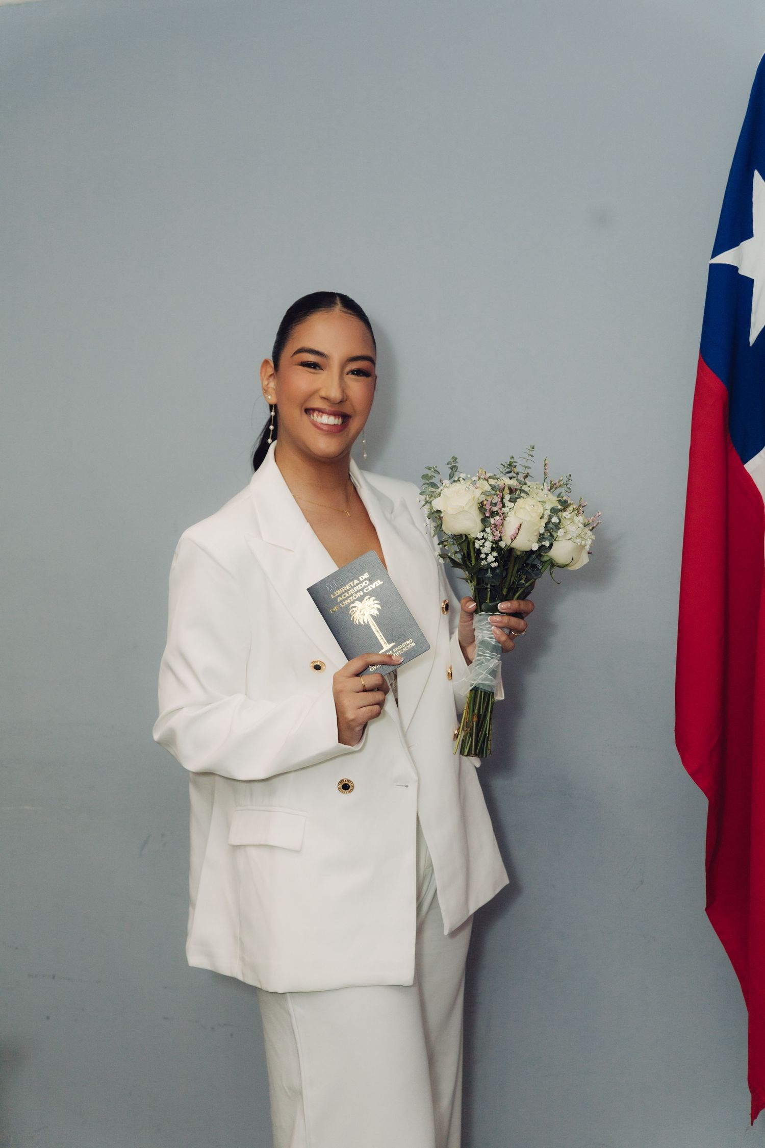 Happy bride in white suit holding bouquet and document beside Chilean flag.