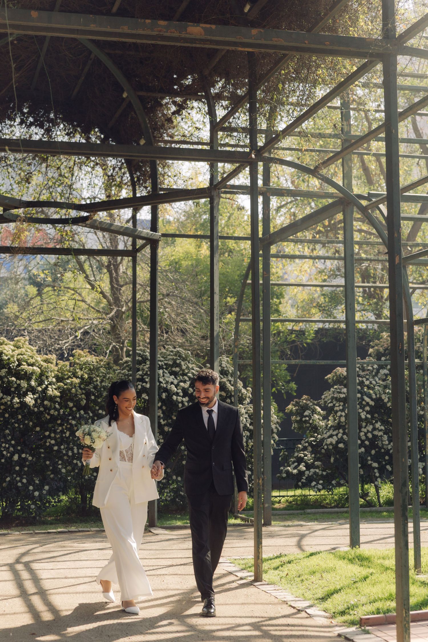 Newlywed couple walking hand in hand through sunlit garden pergola.