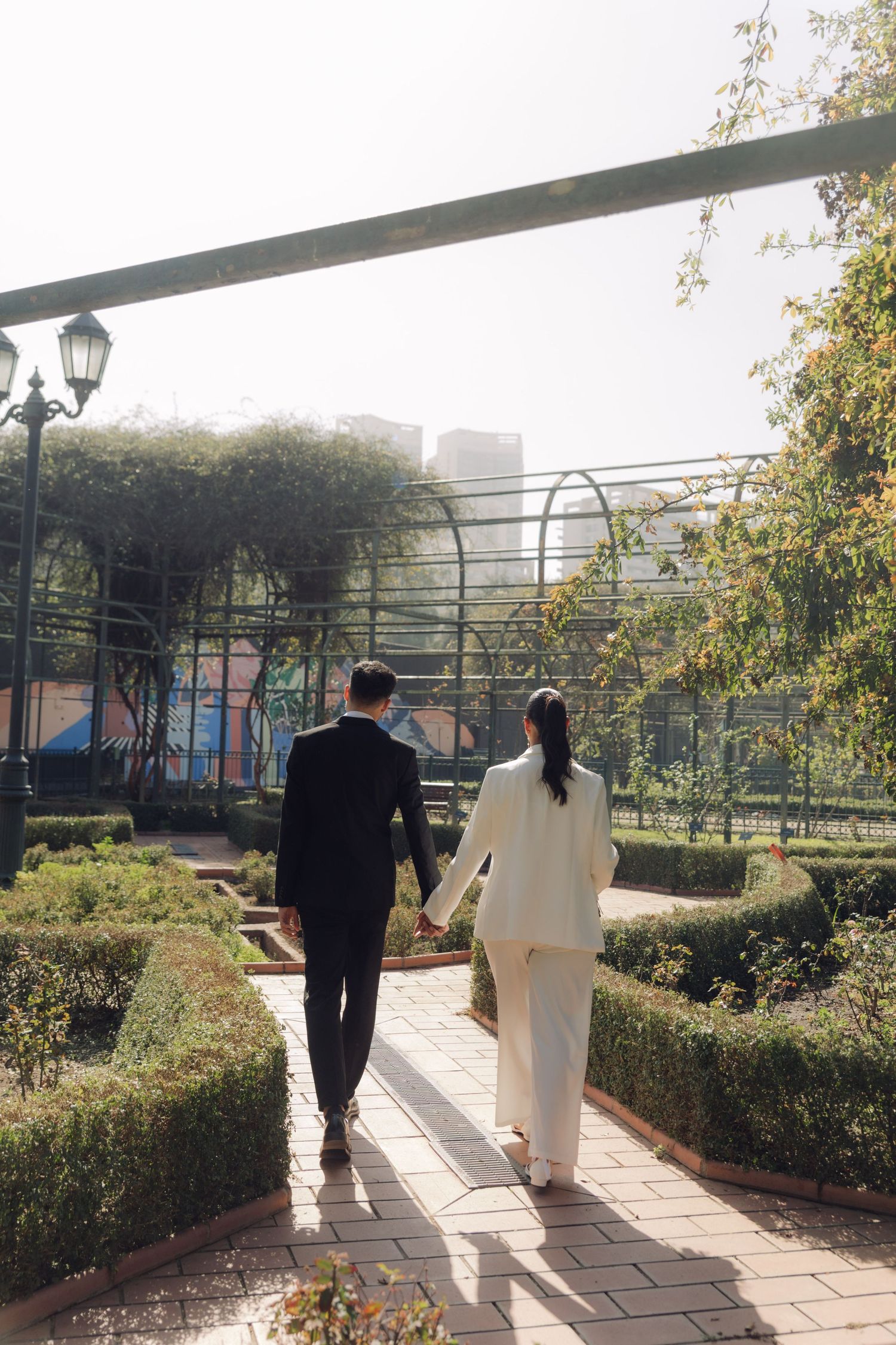 Bride and groom walking away through formal garden path holding hands.