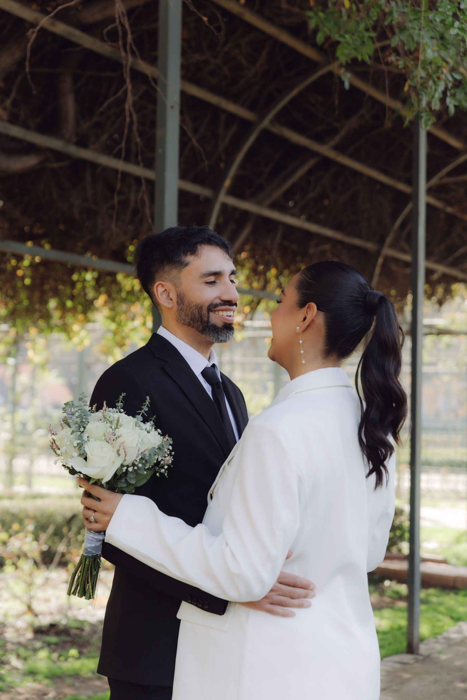Smiling groom in black suit embracing bride in white blazer holding bouquet.