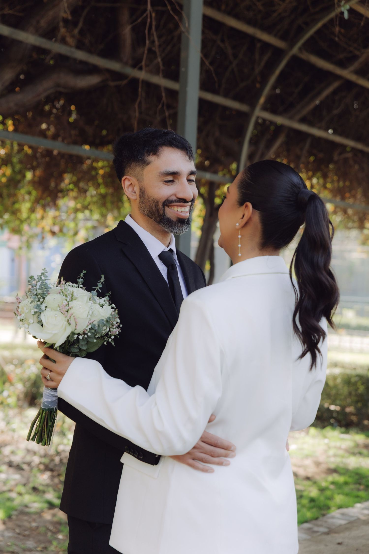 Happy couple embracing under garden pergola, groom in black suit holding white bouquet.
