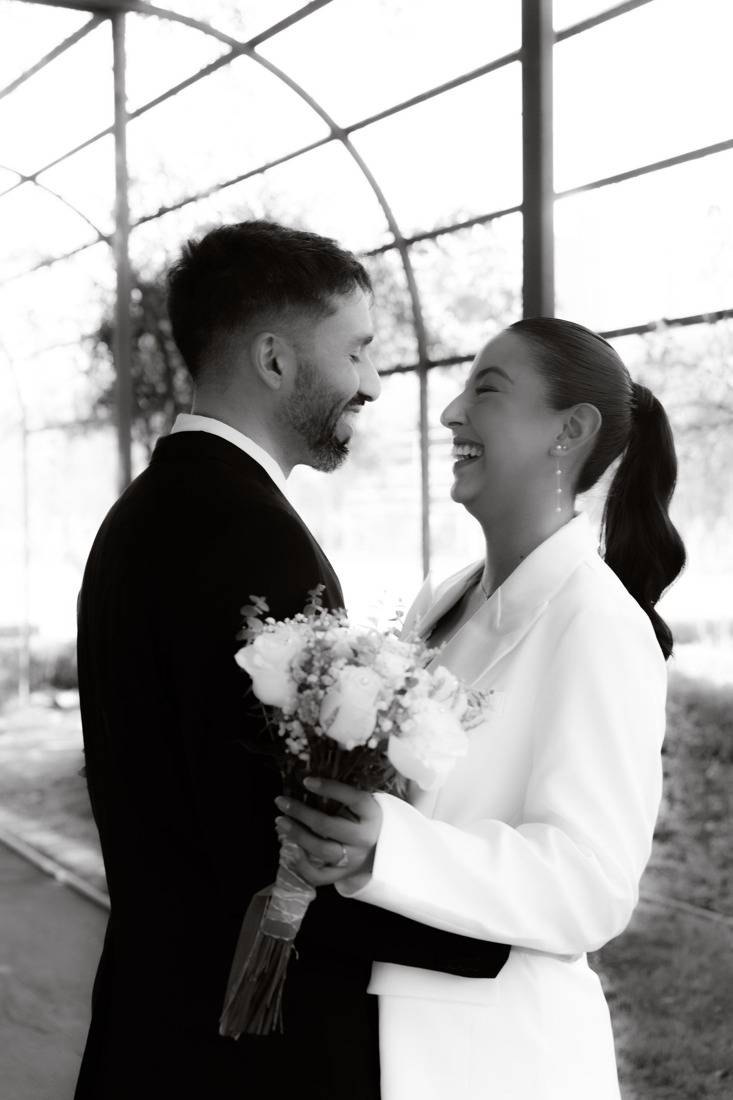Black and white photo of laughing bride and groom holding bouquet under pergola.