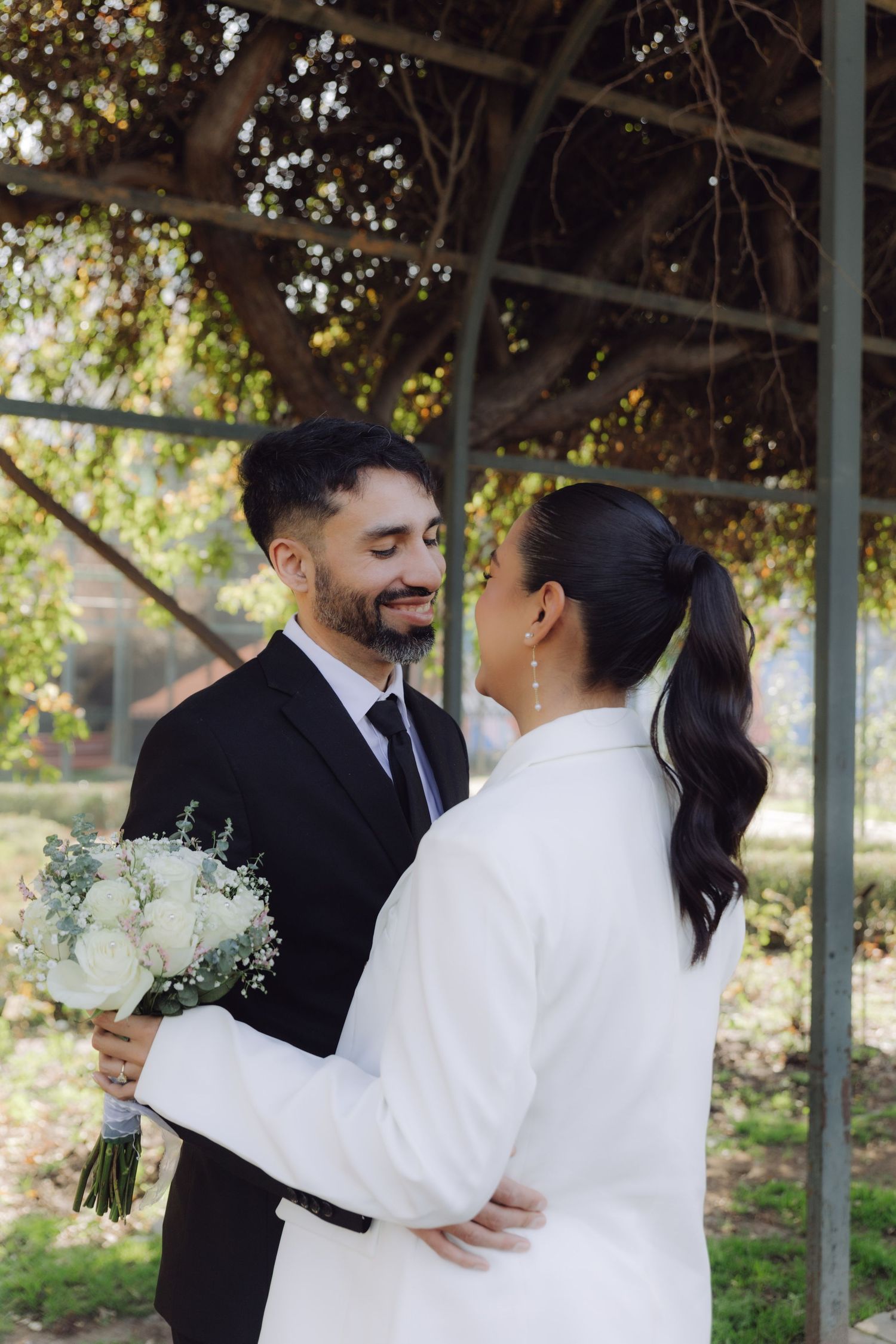 Bearded groom in black suit smiling at bride in white blazer holding bouquet.