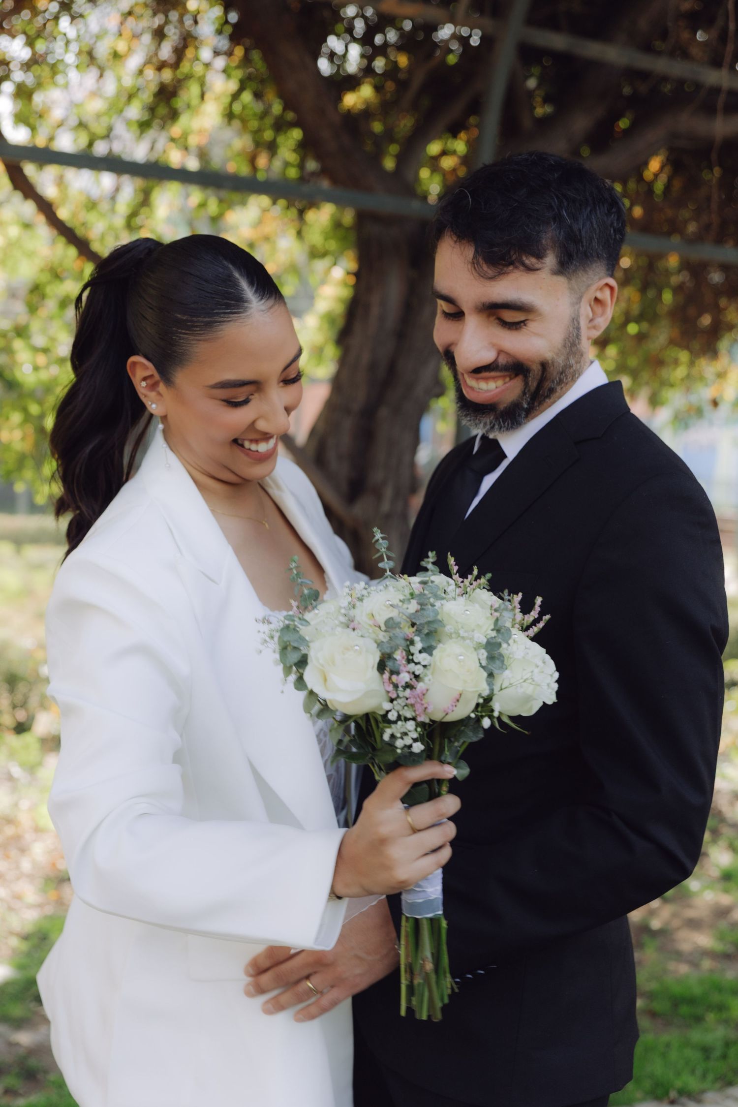 Smiling newlyweds looking at white rose bouquet together under garden tree.