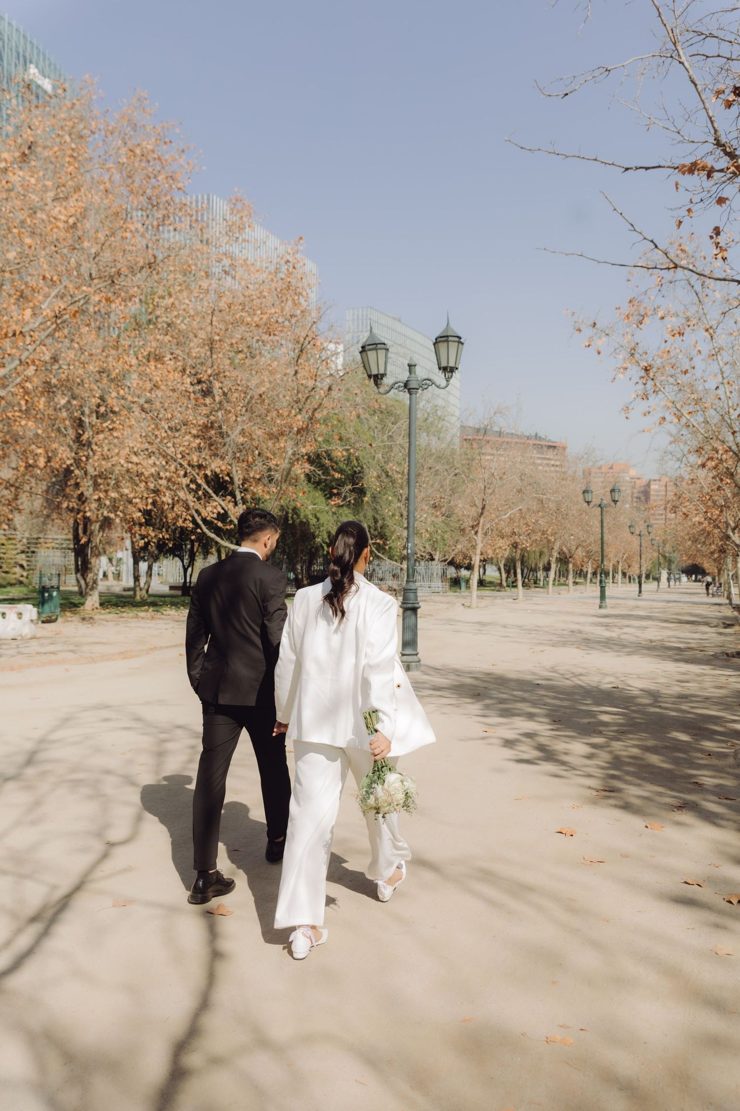 Bride in white suit and groom in black suit walking away in autumn park holding hands.