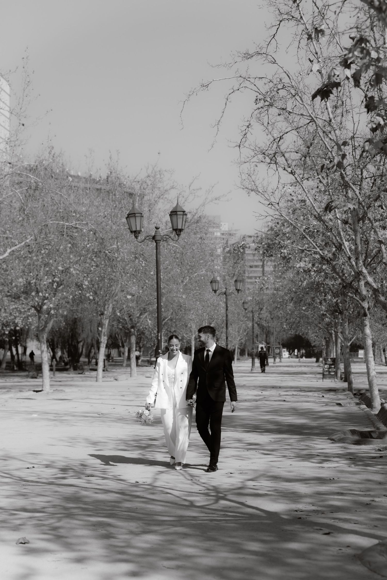 Black and white photo of newlyweds walking hand in hand through a tree-lined park path.