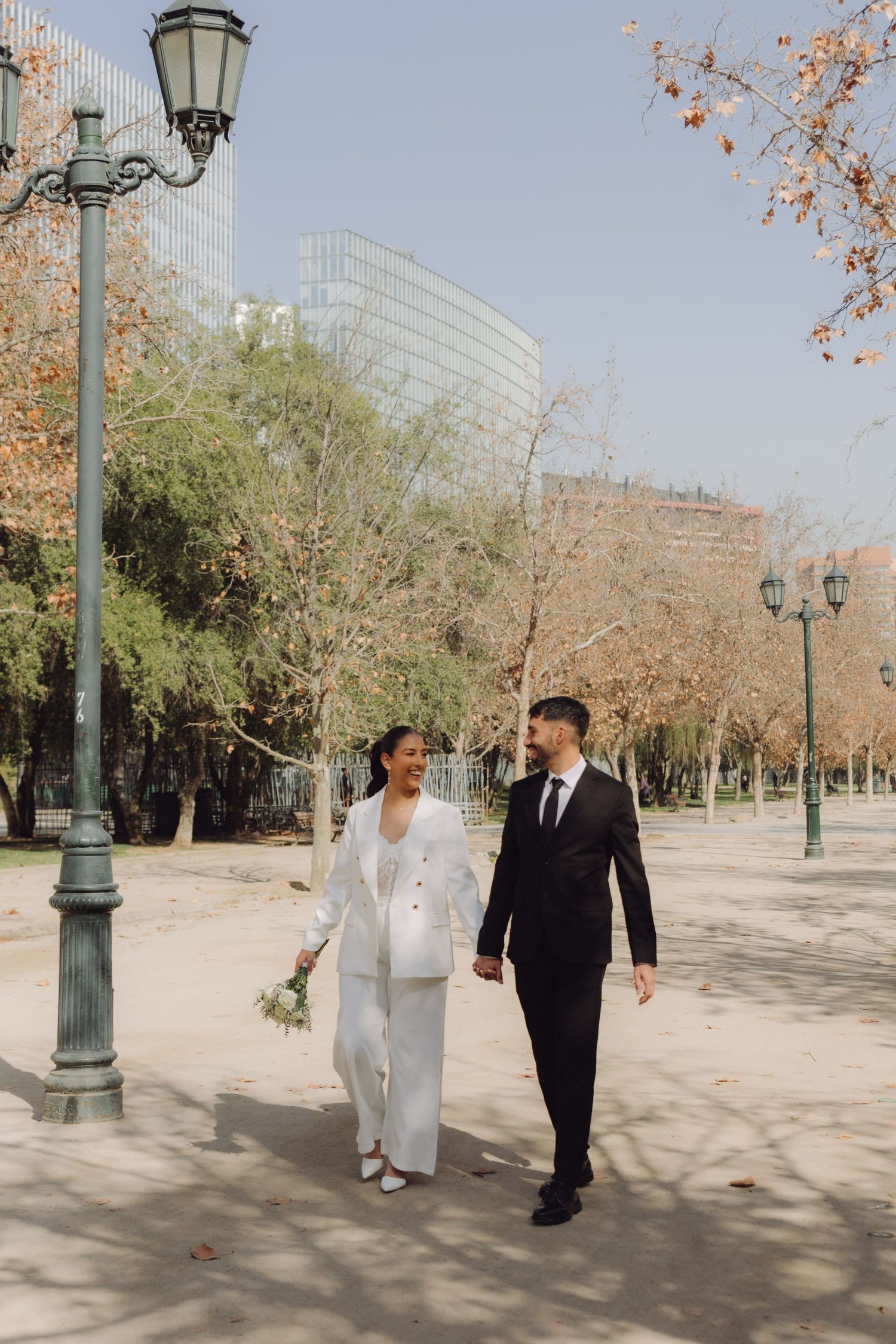 Smiling bride in white pantsuit and groom in black suit walking hand in hand in sunny park.