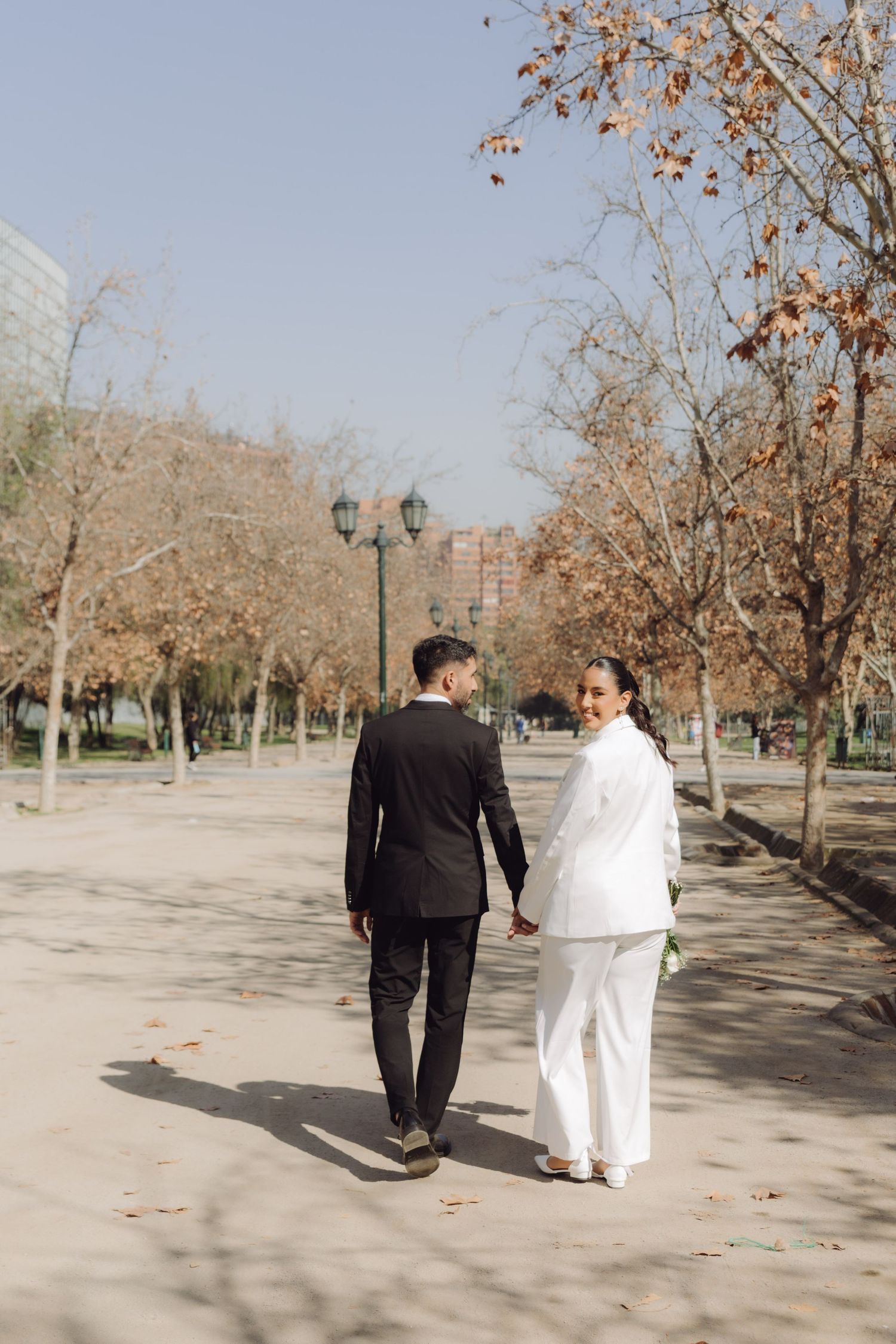 Bride in white suit and groom in black suit walking hand in hand through autumn park path.