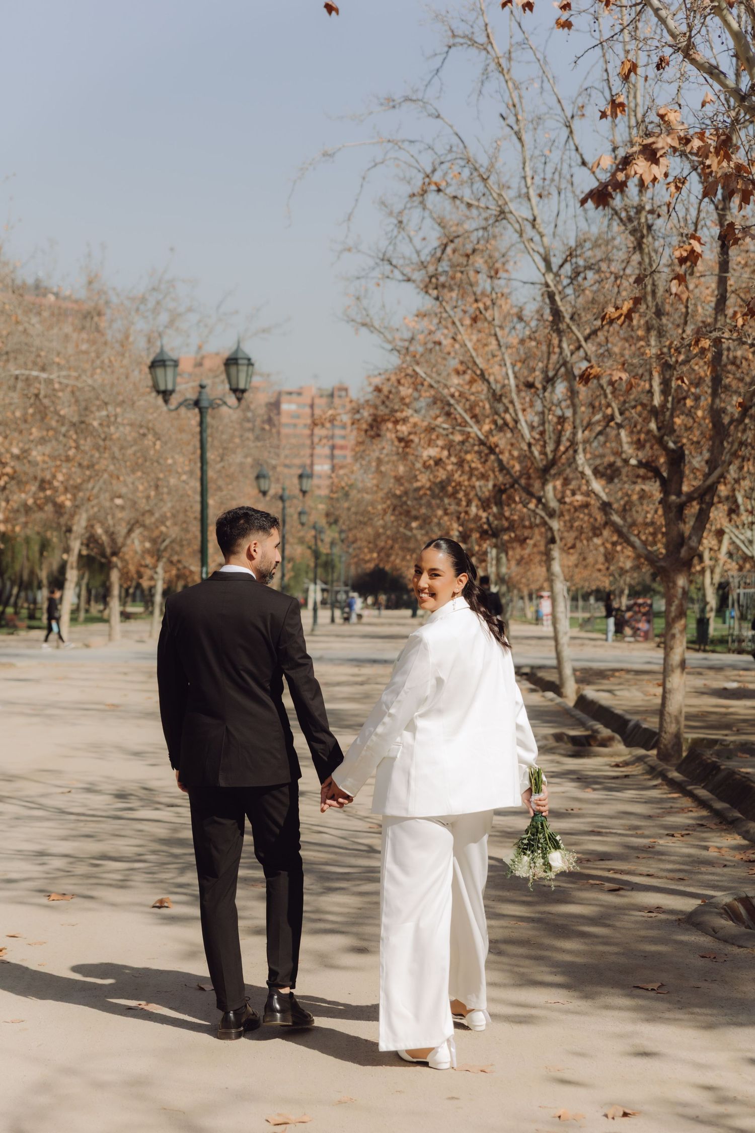 Bride in white blazer holding bouquet smiles back at groom while walking in autumn park.
