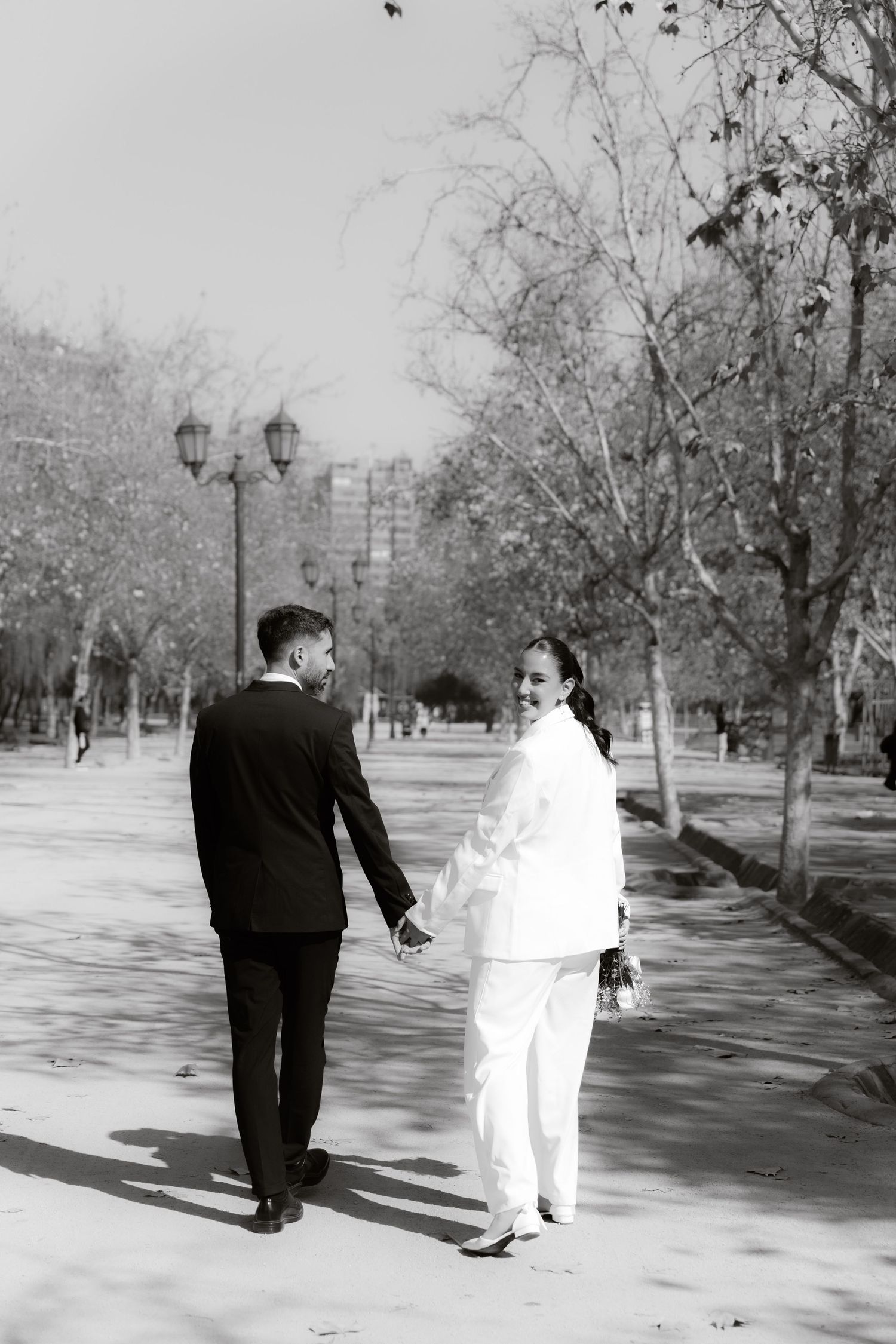 Black and white photo of bride smiling back at groom while walking hand in hand in park.