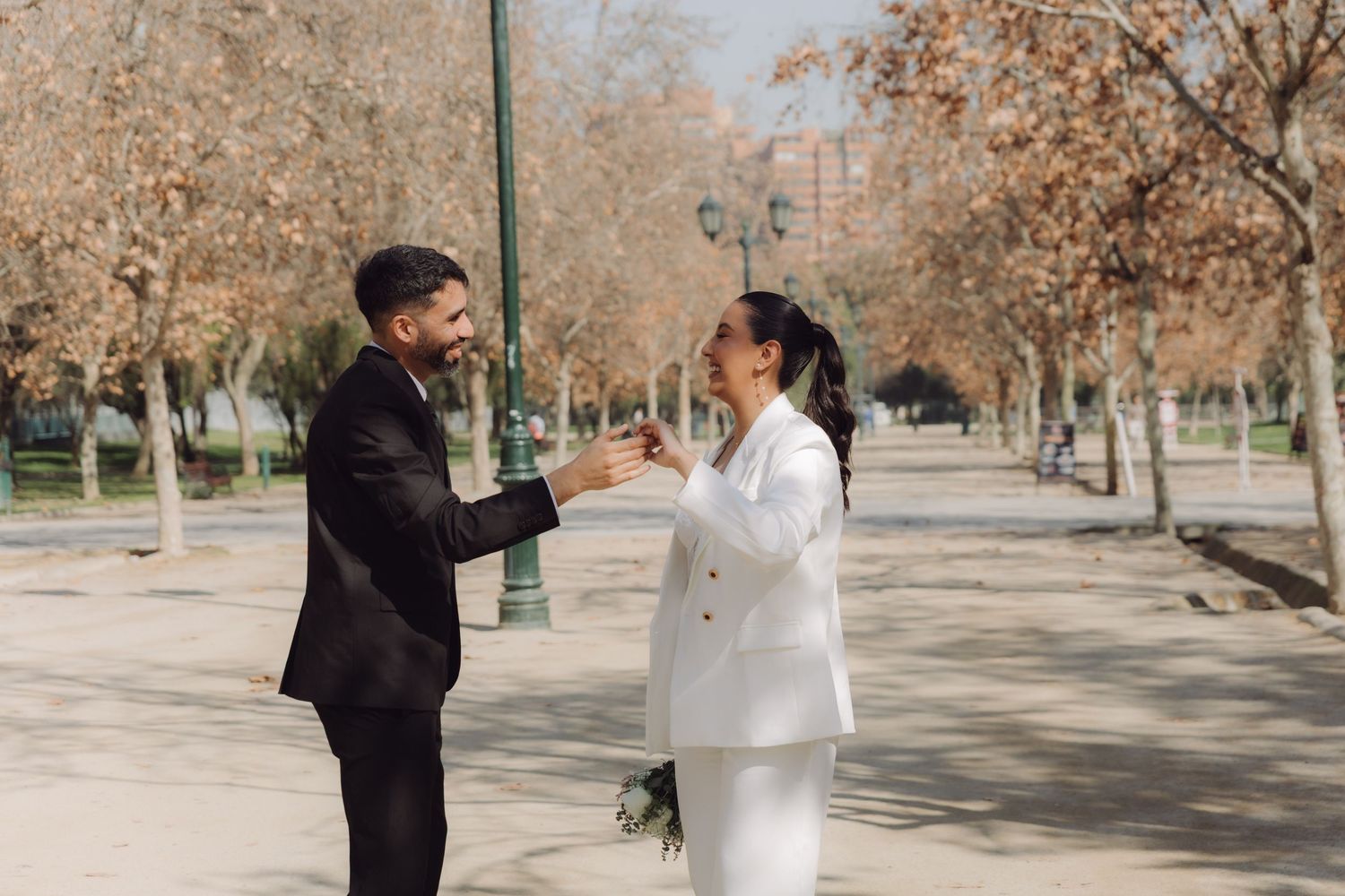Bride and groom in white and black suits laughing and holding hands in a sunlit autumn park.