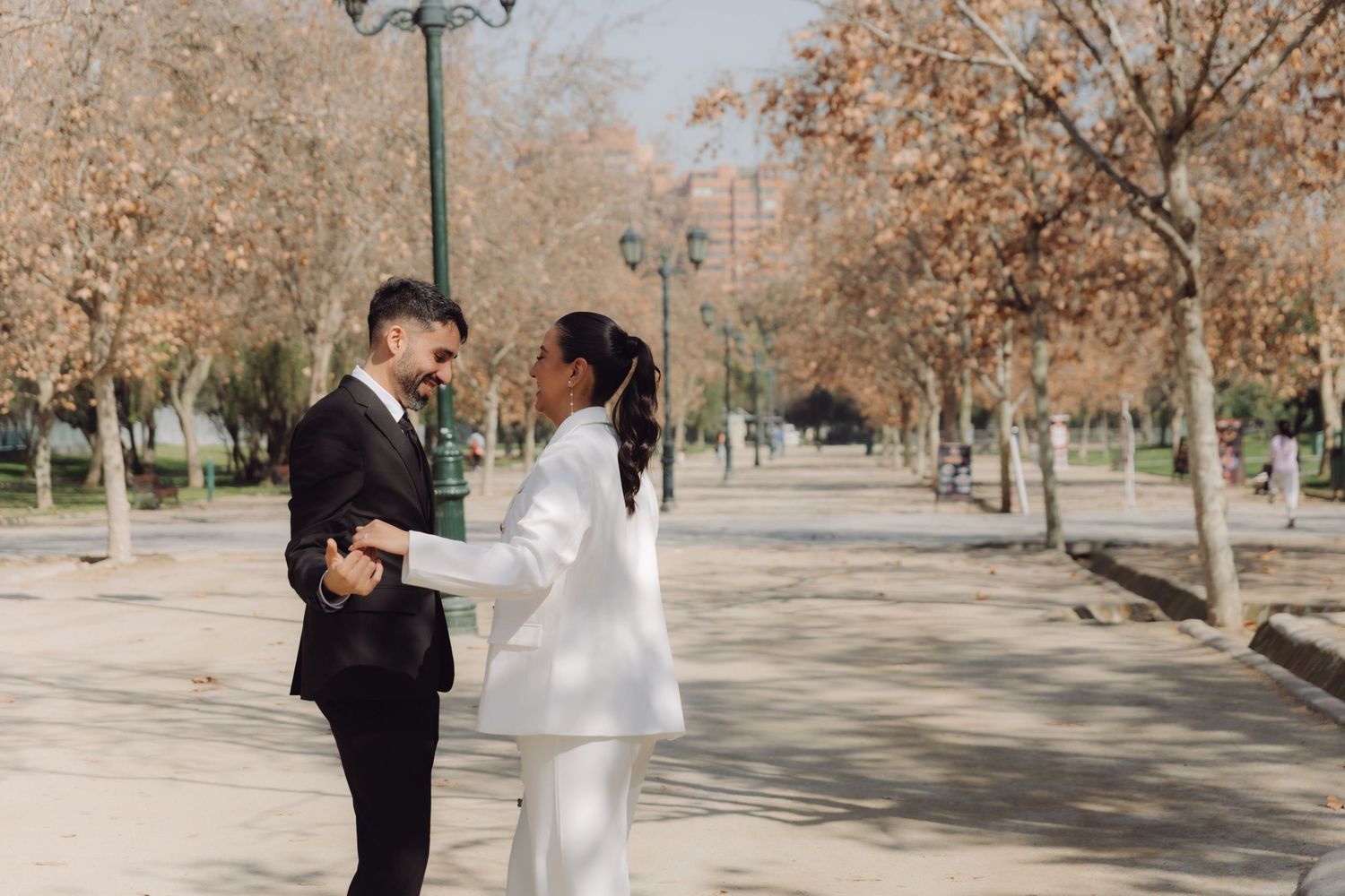Bride in white pantsuit and groom in black suit holding hands facing each other in autumn park.