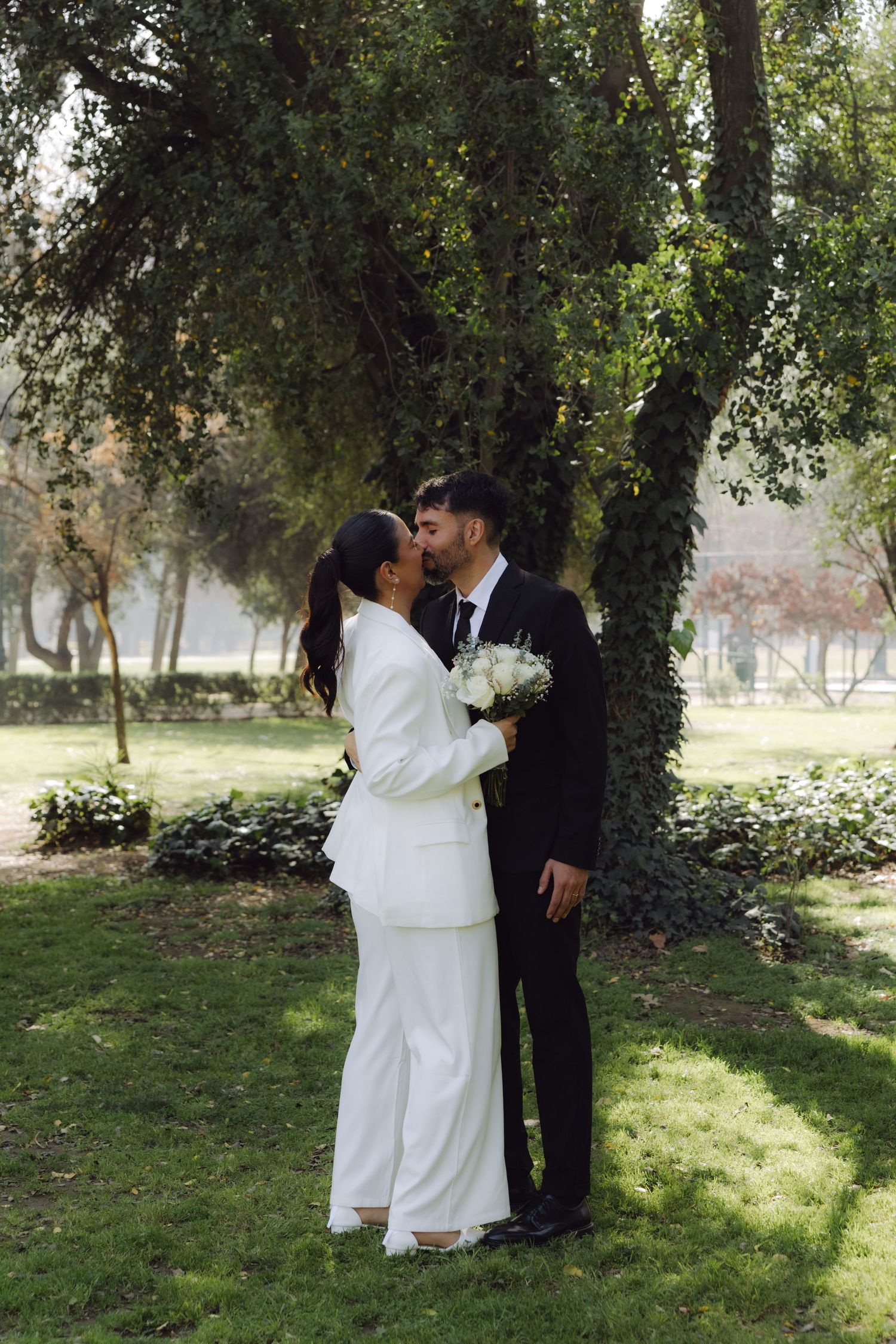 Bride in white suit holding bouquet about to kiss groom under large tree in lush green park.
