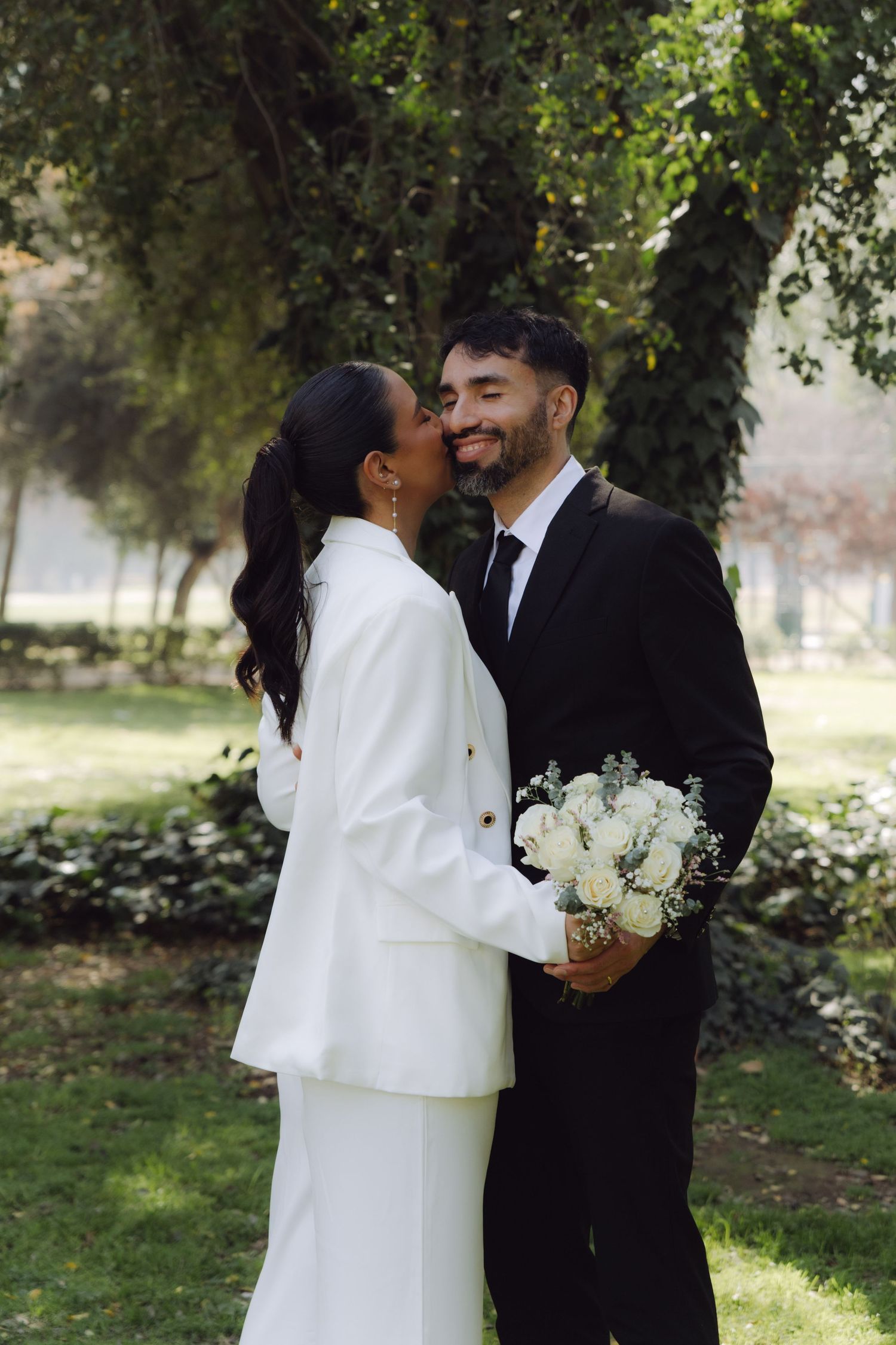 Bride in white suit kissing groom on cheek as he smiles holding bouquet in green park.