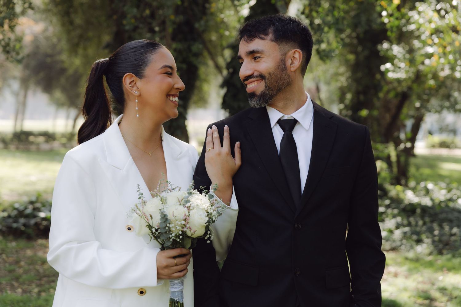 Smiling bride and groom in white blazer and black suit walk together holding white flower bouquet in park.
