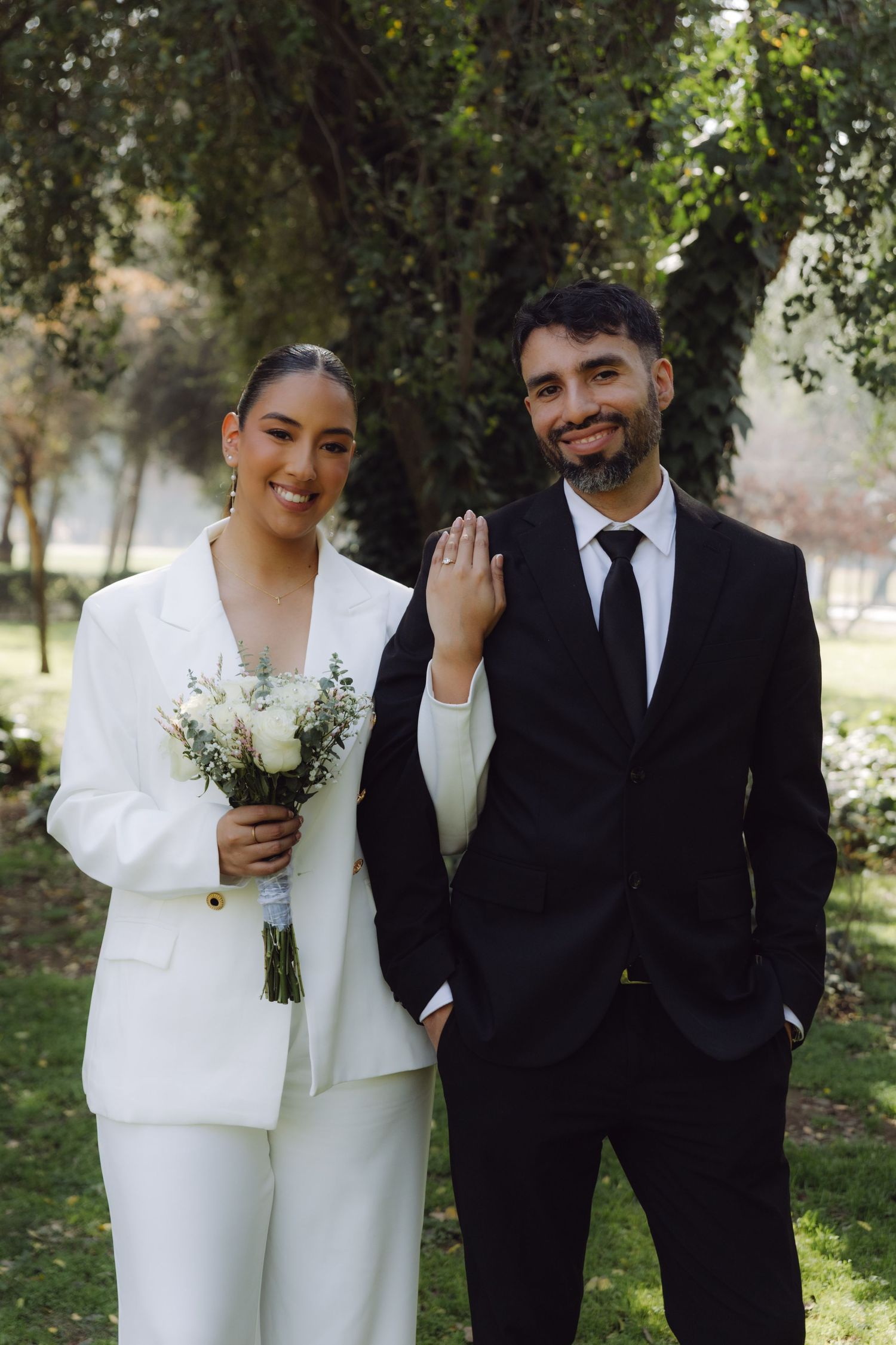 Happy bride in white pantsuit holds bouquet and shows ring beside groom in black suit in sunny park.