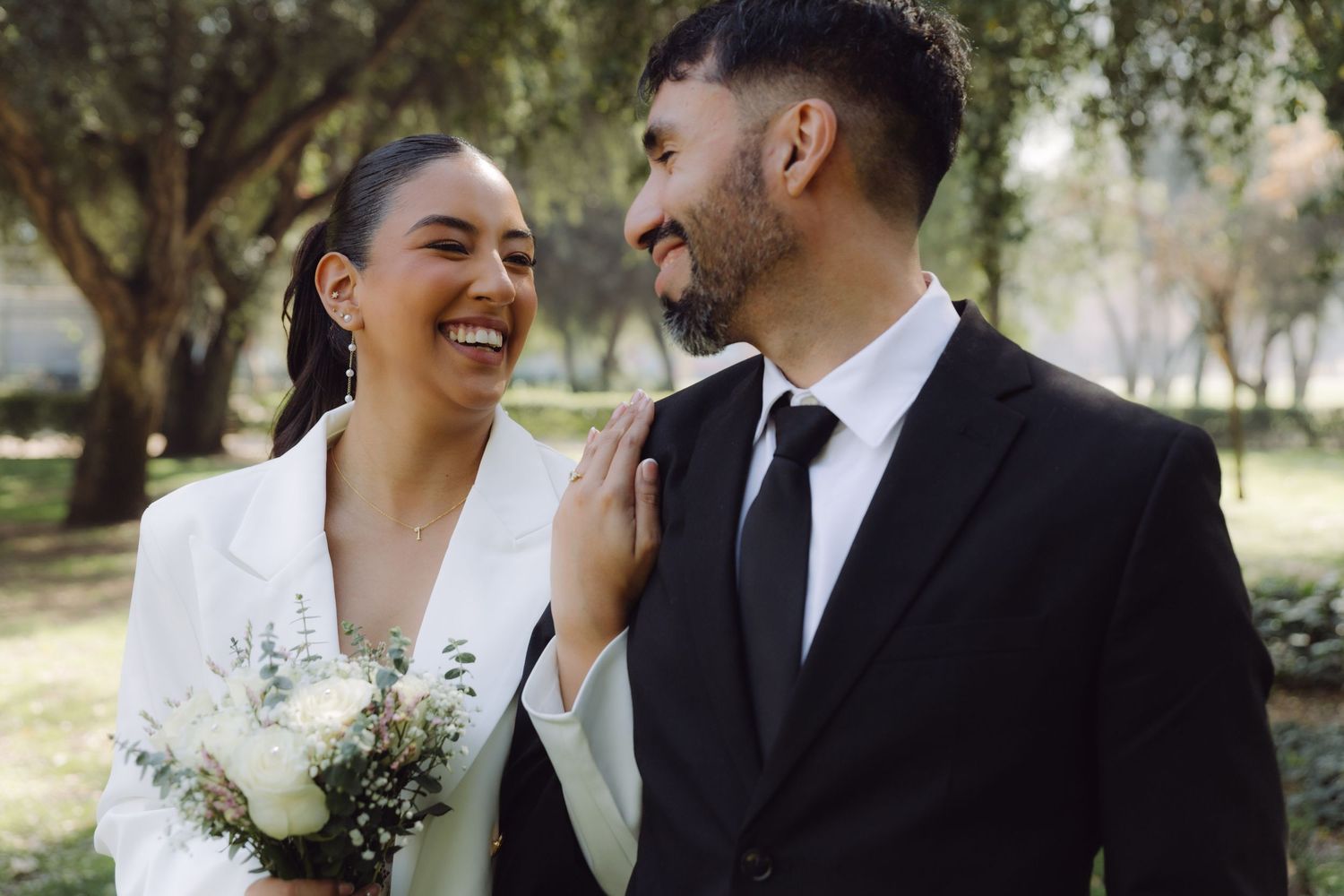 Joyful bride in white blazer and groom in black suit smile at each other holding white bouquet outdoors.