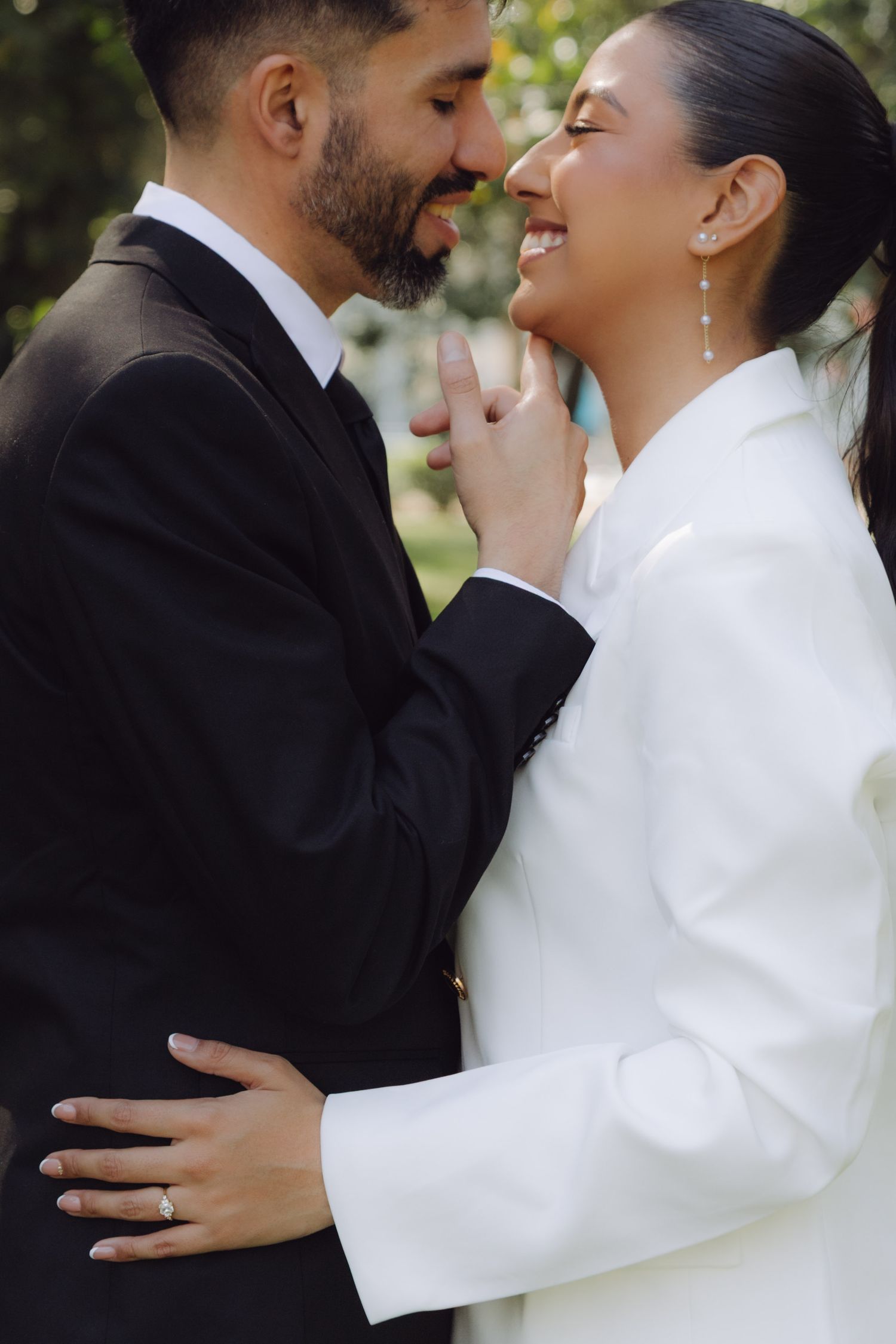 Laughing bride in white blazer and groom in black suit share intimate moment touching foreheads in park.