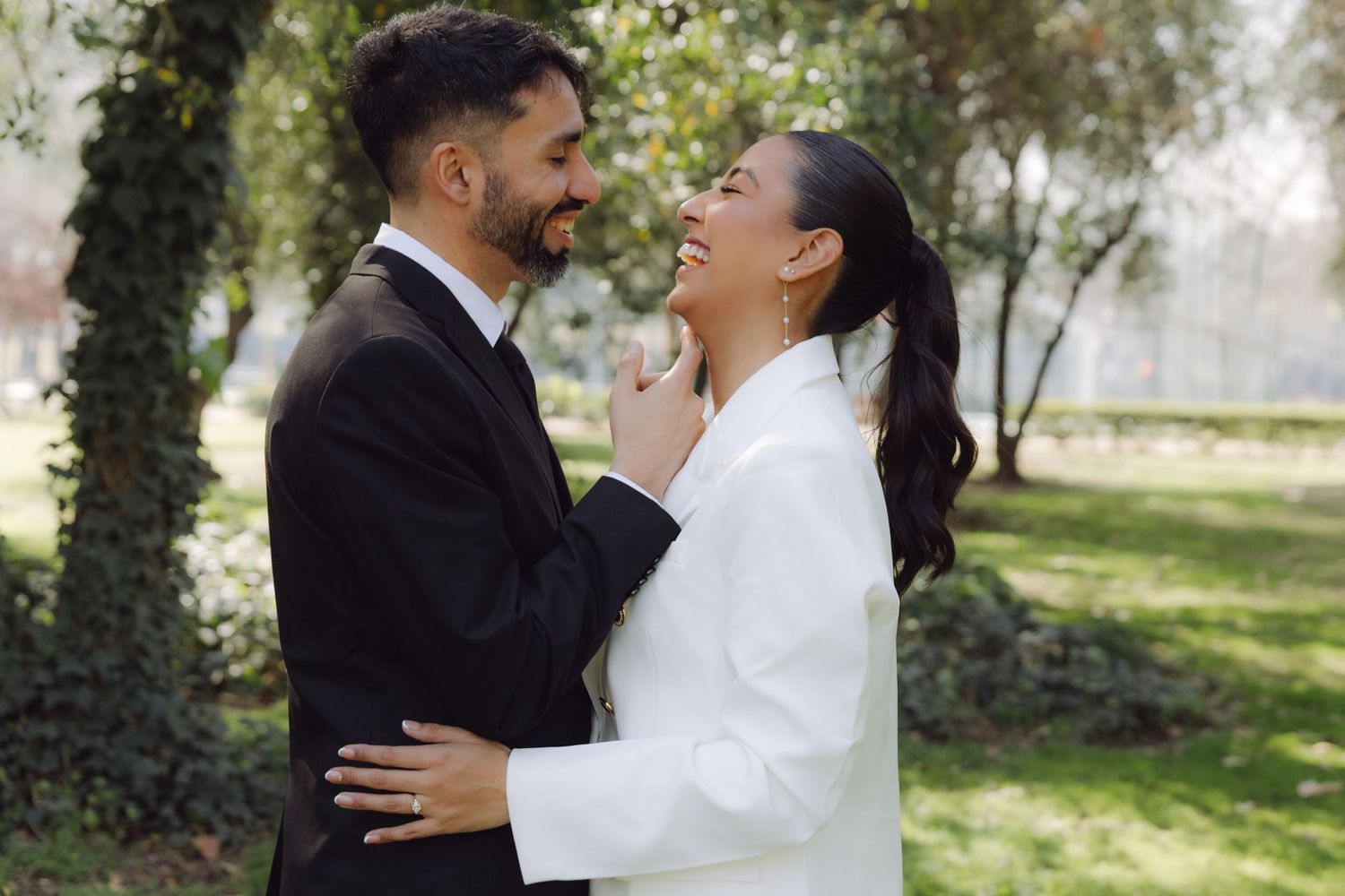 Bride in white blazer and groom in black suit embrace and laugh together in sunlit garden setting.