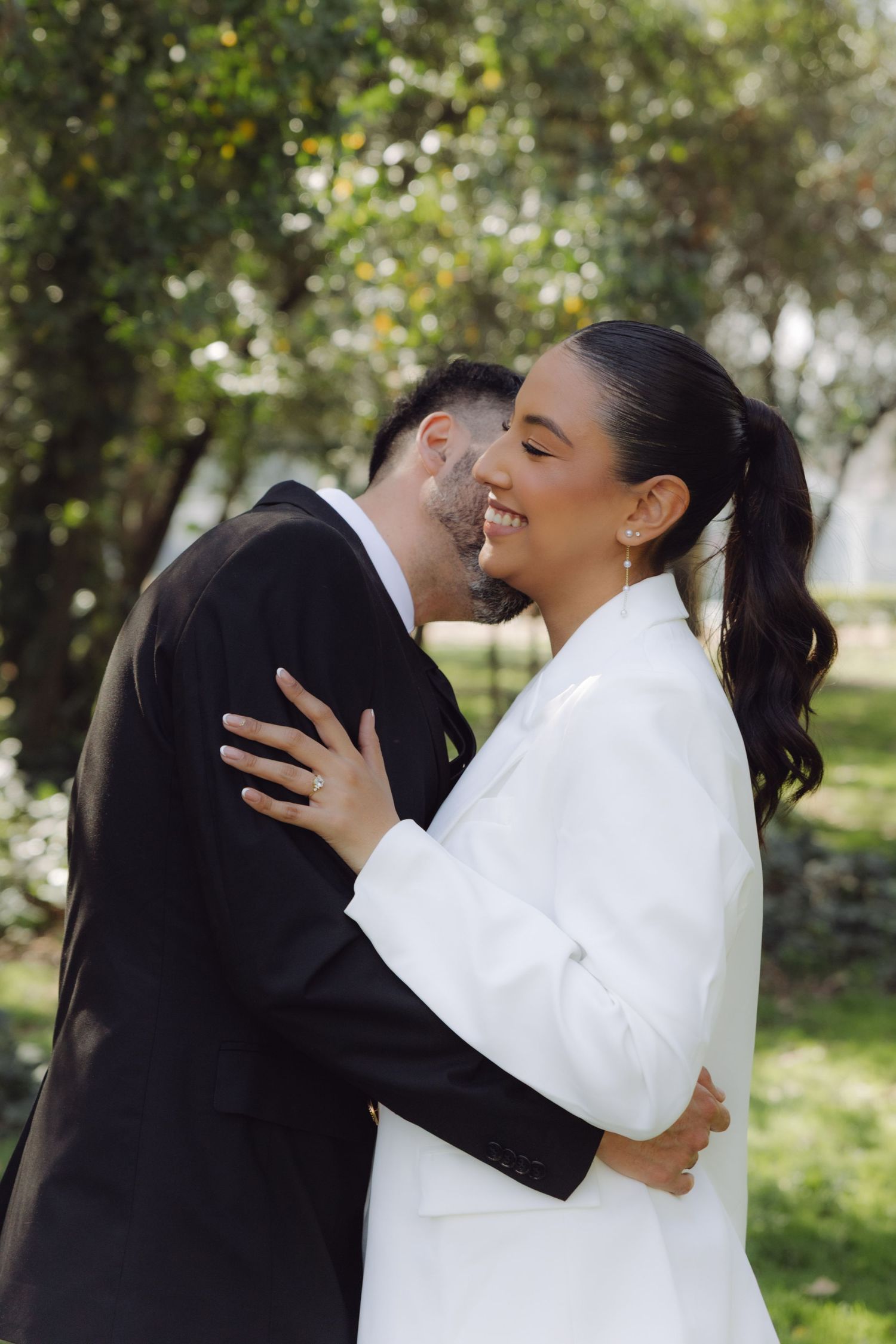 Smiling bride in white blazer embraces groom in black suit during outdoor wedding photo session in green park.