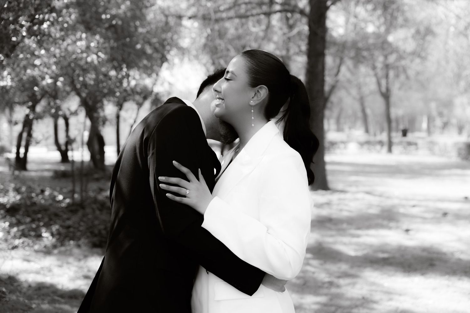 Black and white photo of bride and groom sharing a tender kiss in a tree-lined park setting.