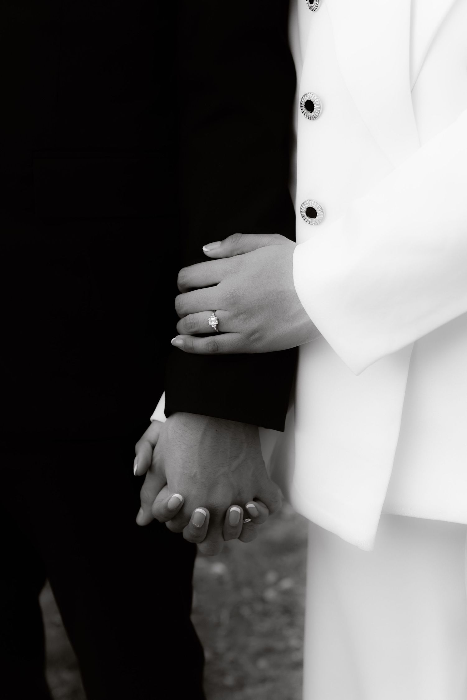 Close-up black and white photo of bride and groom holding hands, showcasing diamond engagement ring.
