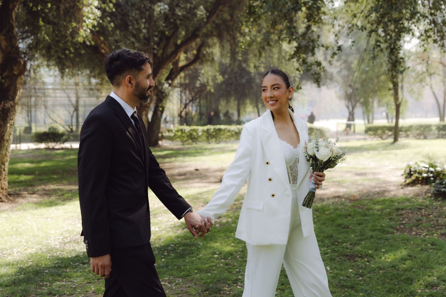 Joyful bride in white pantsuit holding bouquet walks hand in hand with groom in sunny park.