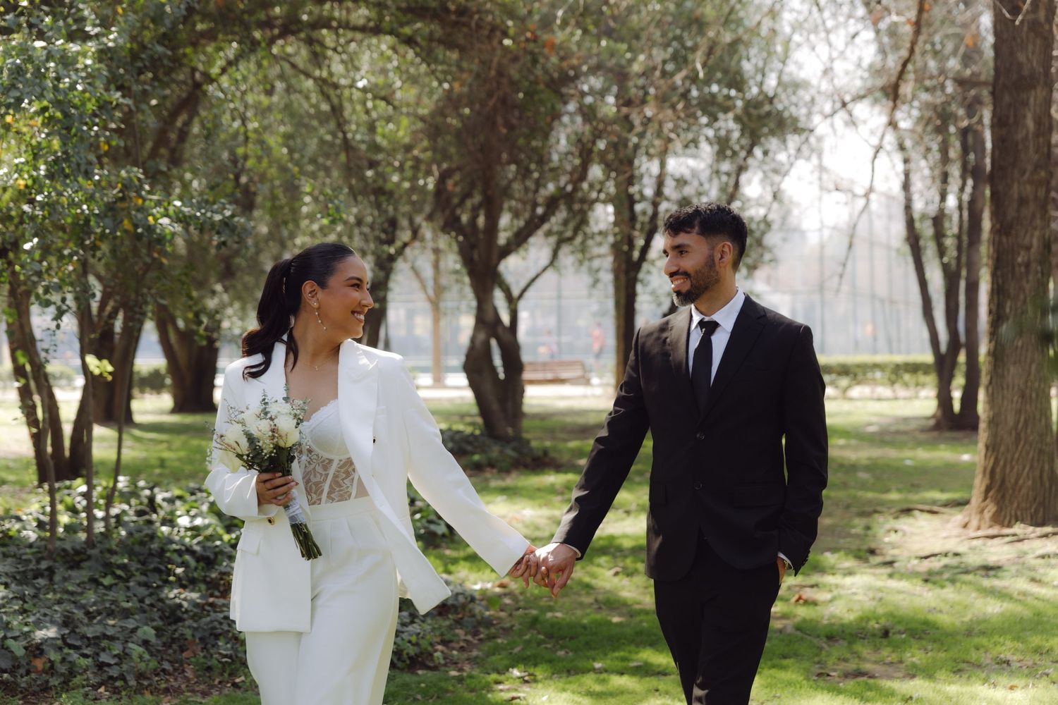 Smiling newlyweds in coordinating black and white suits walk hand in hand through a green park.
