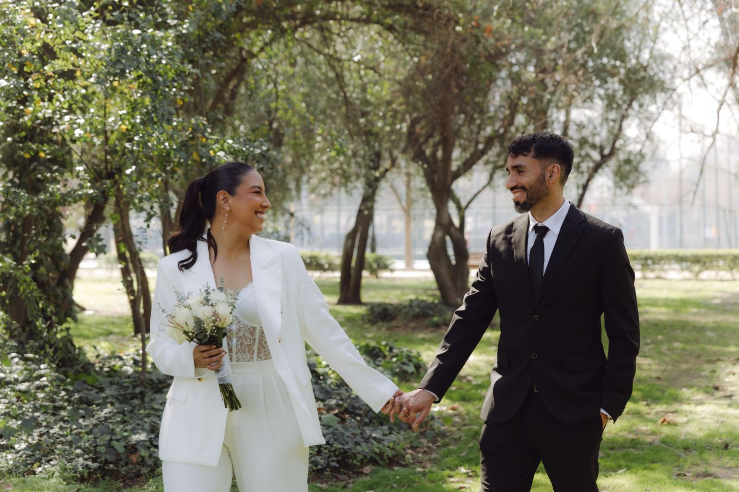 Happy bride and groom in elegant suits hold hands and smile at each other in a sunlit park.