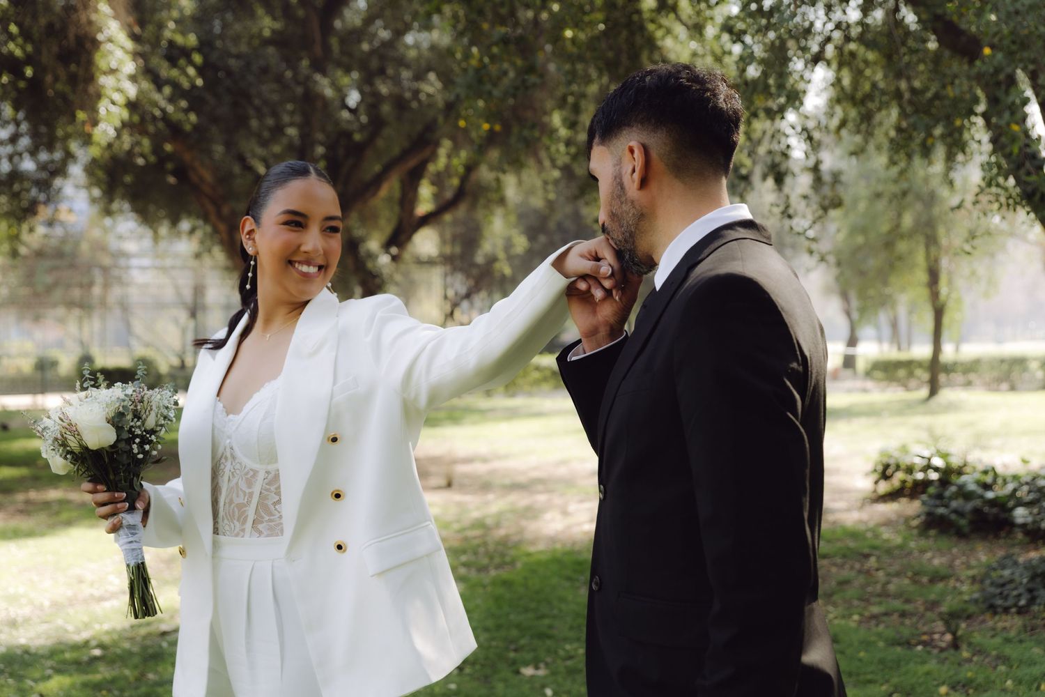 Bride in white blazer extends hand to groom who kisses it while holding bouquet in garden setting.