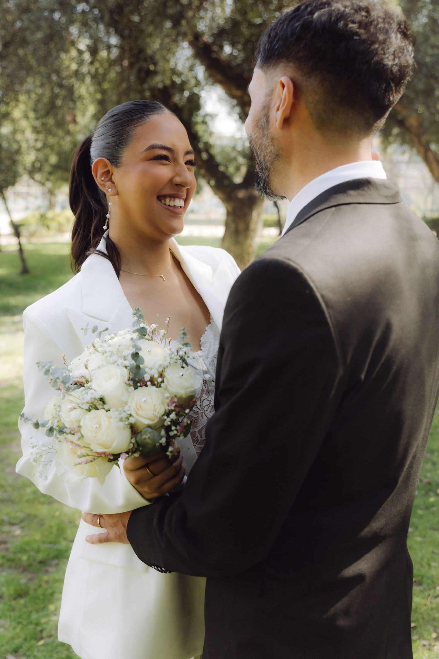 Radiant bride in white blazer holding white bouquet smiles at groom during outdoor wedding portraits.