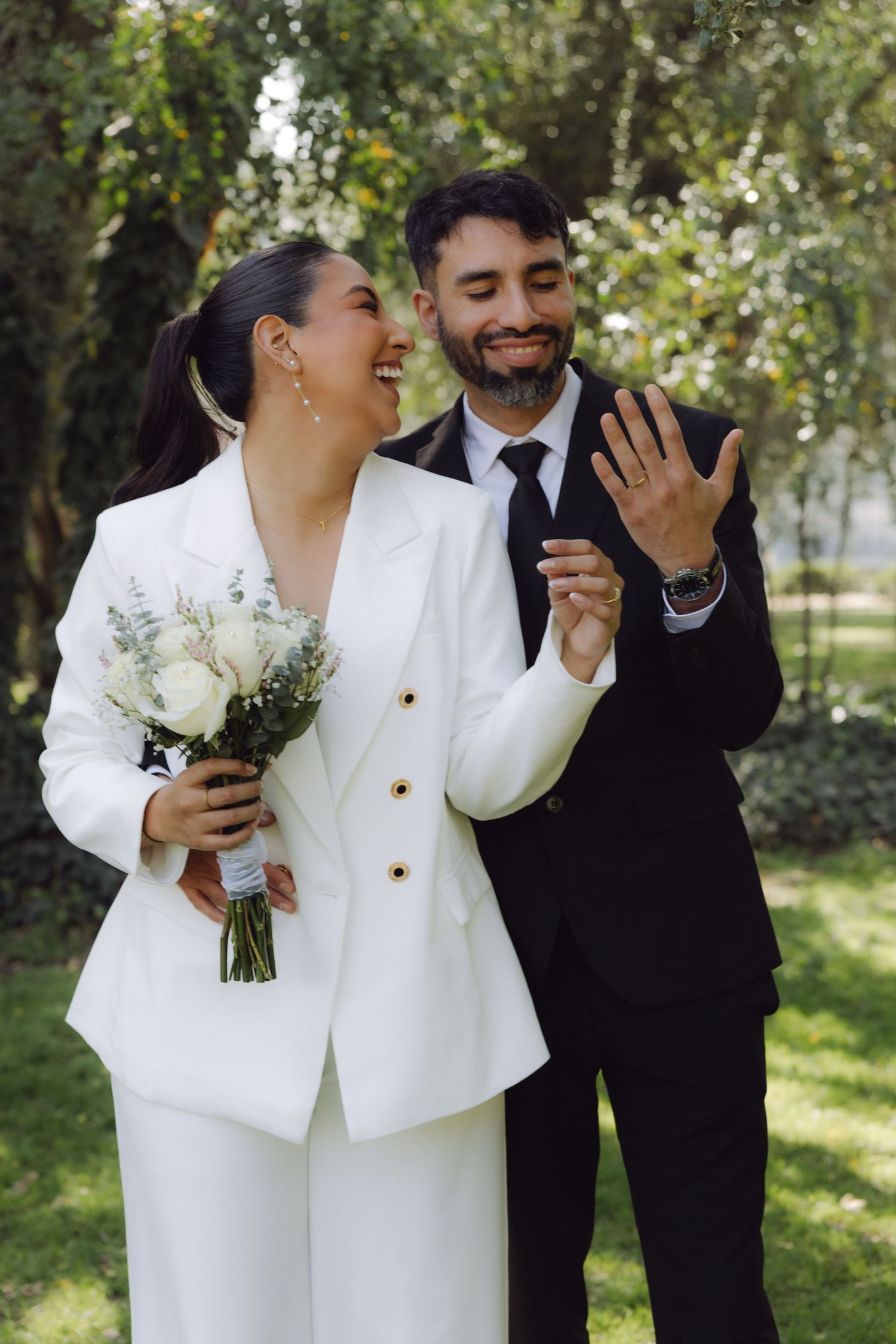 Laughing bride in white suit holding bouquet with groom in black suit showing wedding ring outdoors.