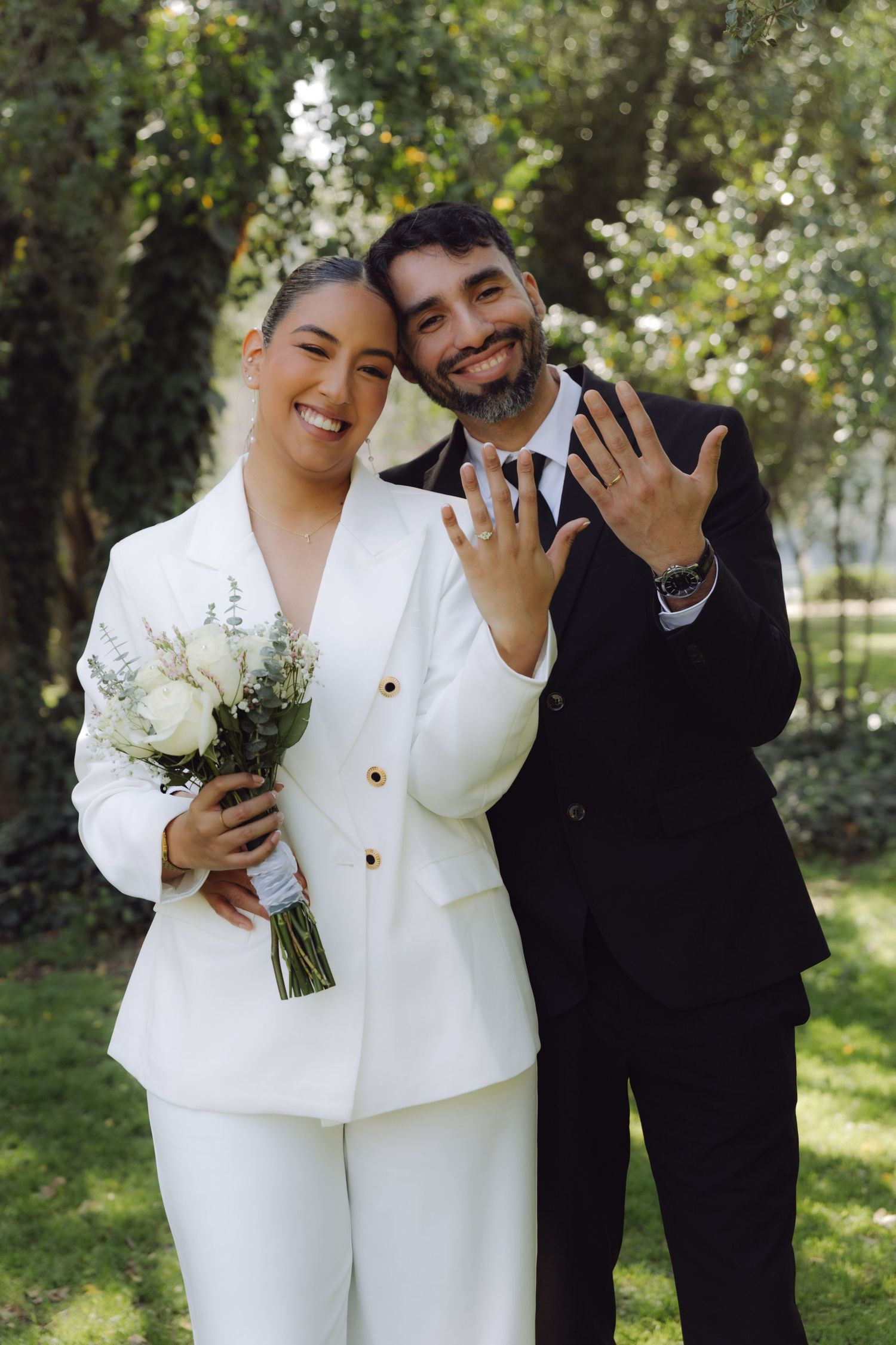 Happy newlyweds posing in park, bride in white suit with bouquet, groom showing wedding ring.