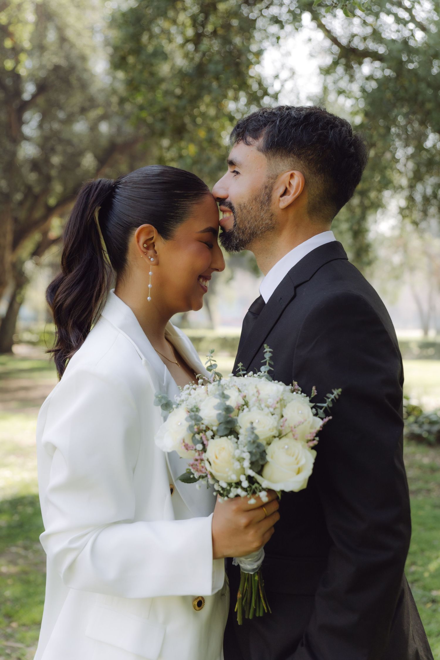 Smiling bride and groom in formal attire sharing intimate moment in green park setting.