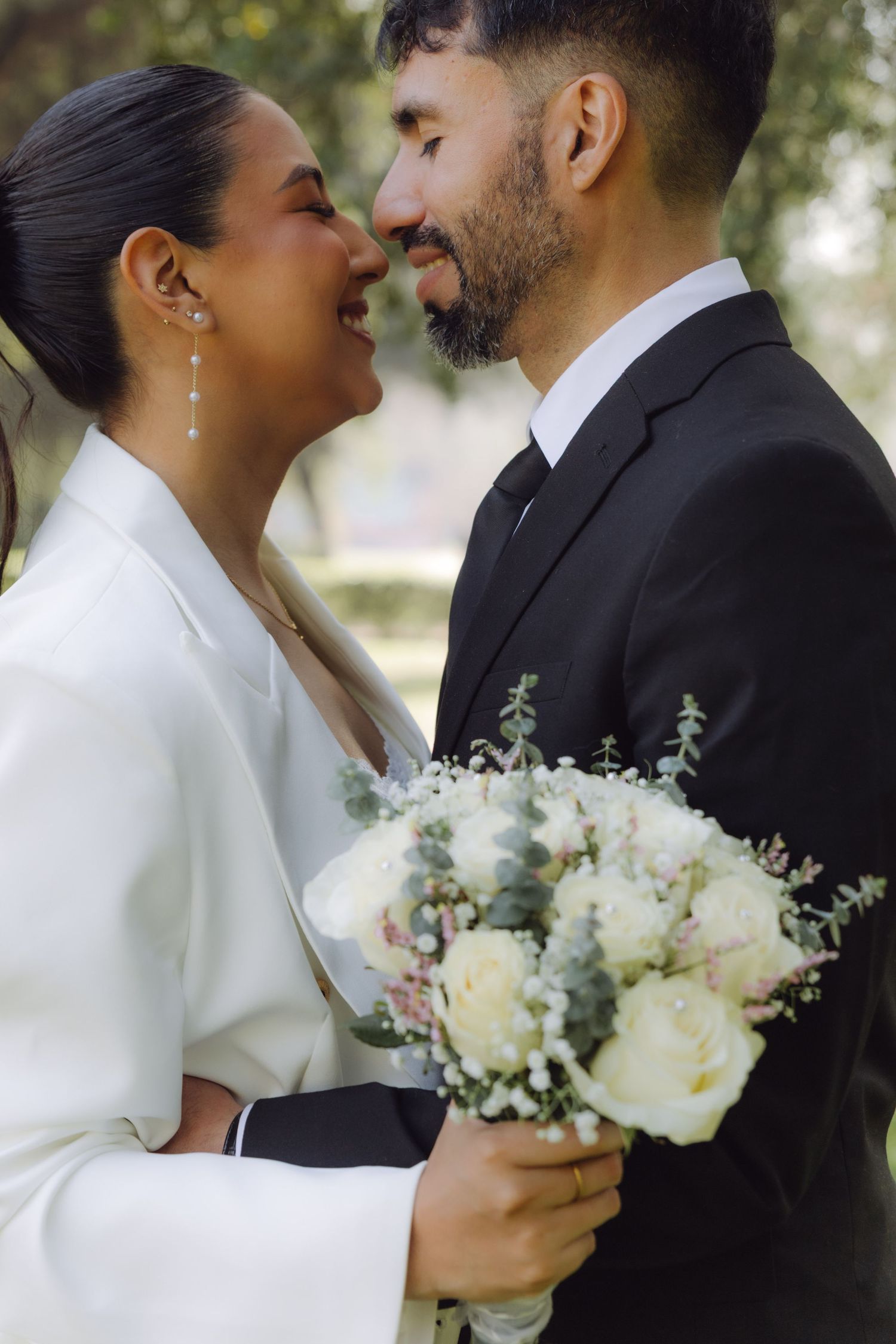 Close-up of newlyweds nearly kissing, bride in white suit holding white rose bouquet outdoors.