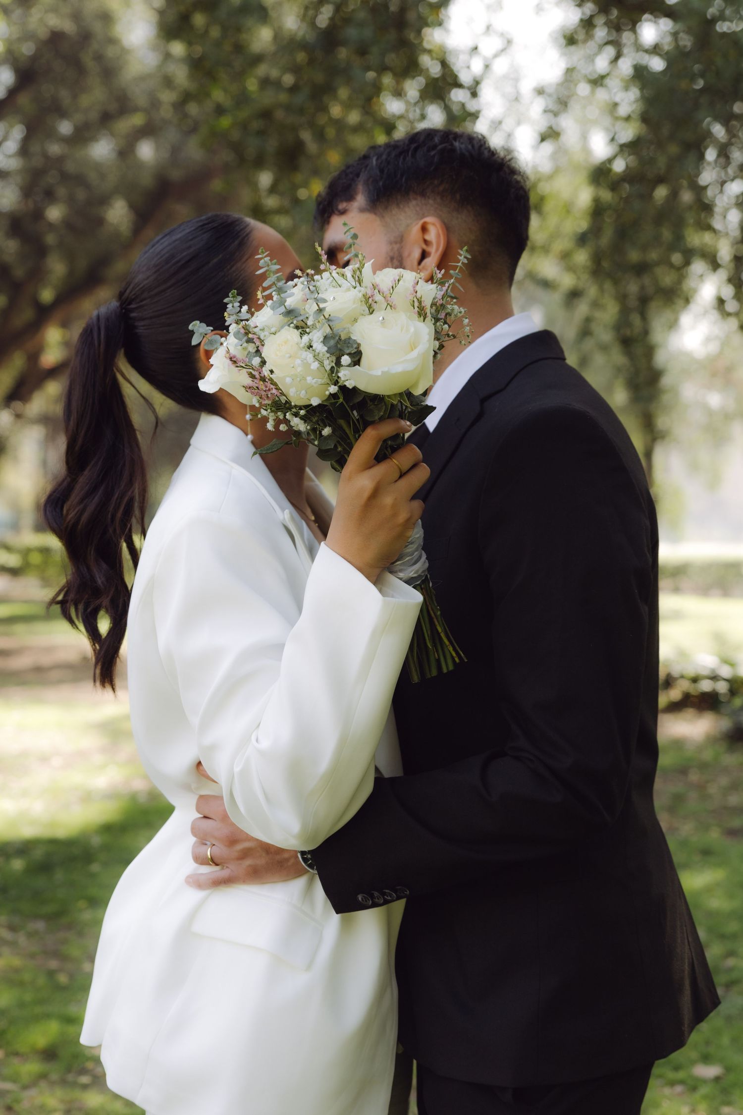 Bride and groom sharing a kiss hidden behind white rose wedding bouquet in park.