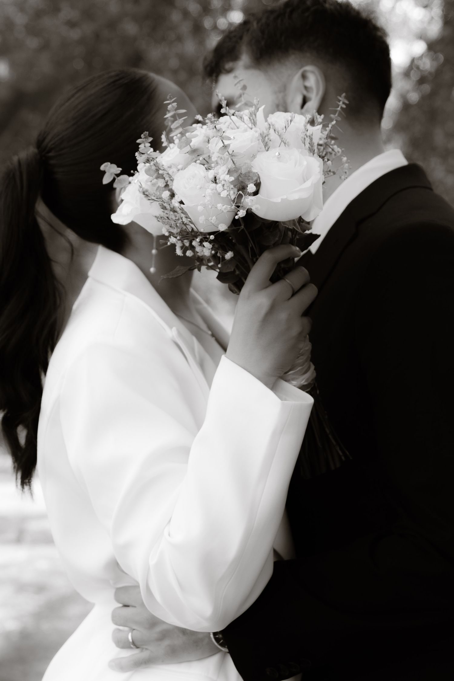 Black and white photo of couple kissing behind bridal bouquet of white roses and greenery.