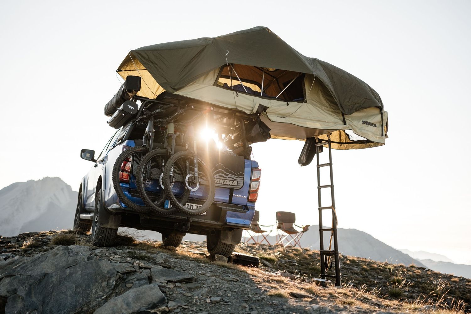 Rooftop tent mounted on SUV overlooks scenic mountain landscape at sunset with camping gear visible.