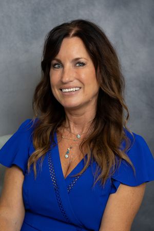 A smiling woman with long brown hair wearing a blue wrap dress and layered necklaces poses for a professional headshot.