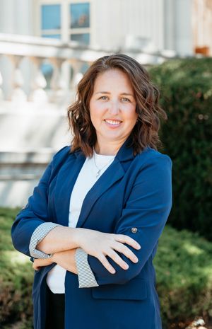 Professional headshot portrait of someone in a navy blazer and white blouse against an outdoor background.