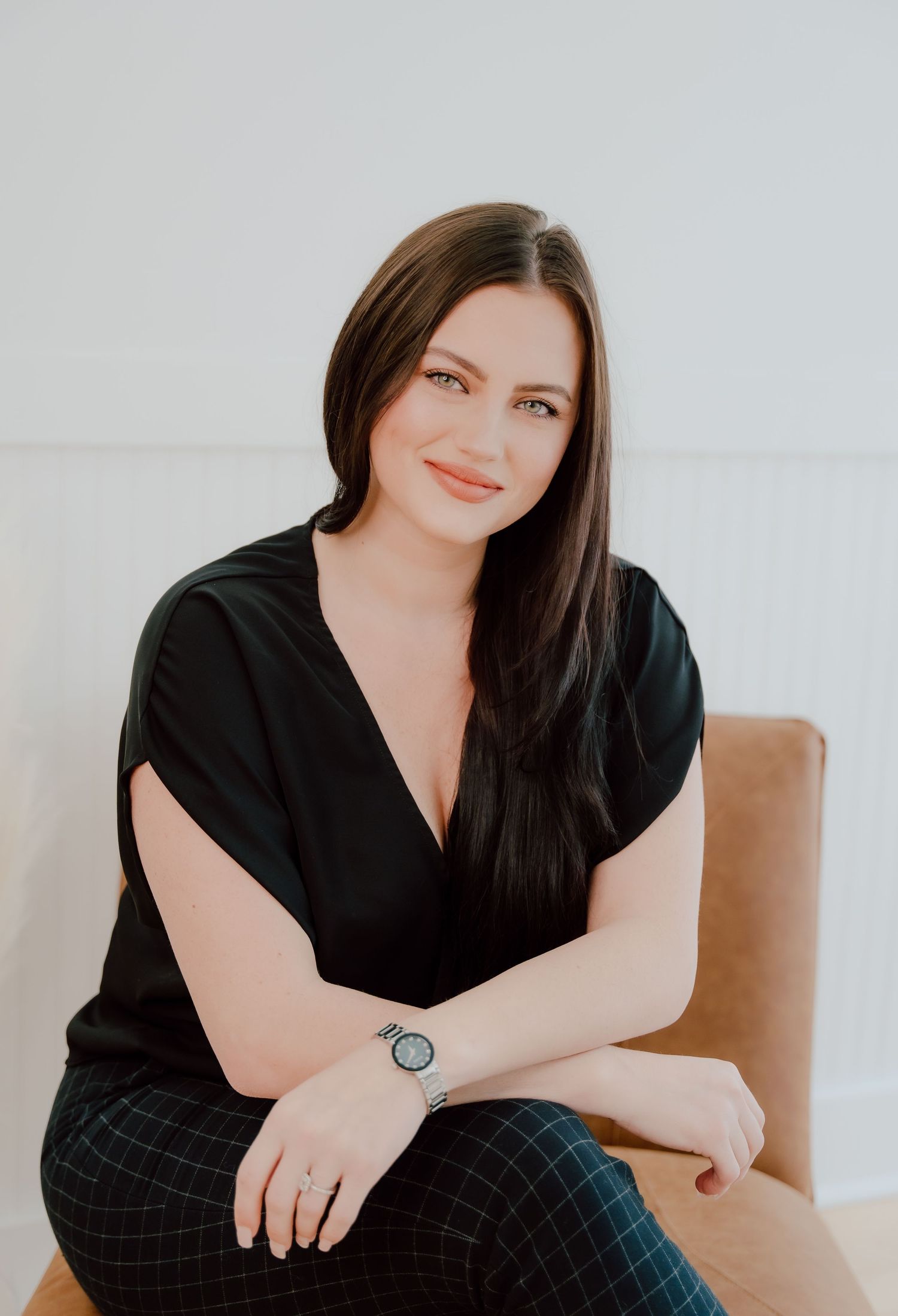 Professional headshots of a person in a black top and plaid pants seated on a brown chair against a light background.